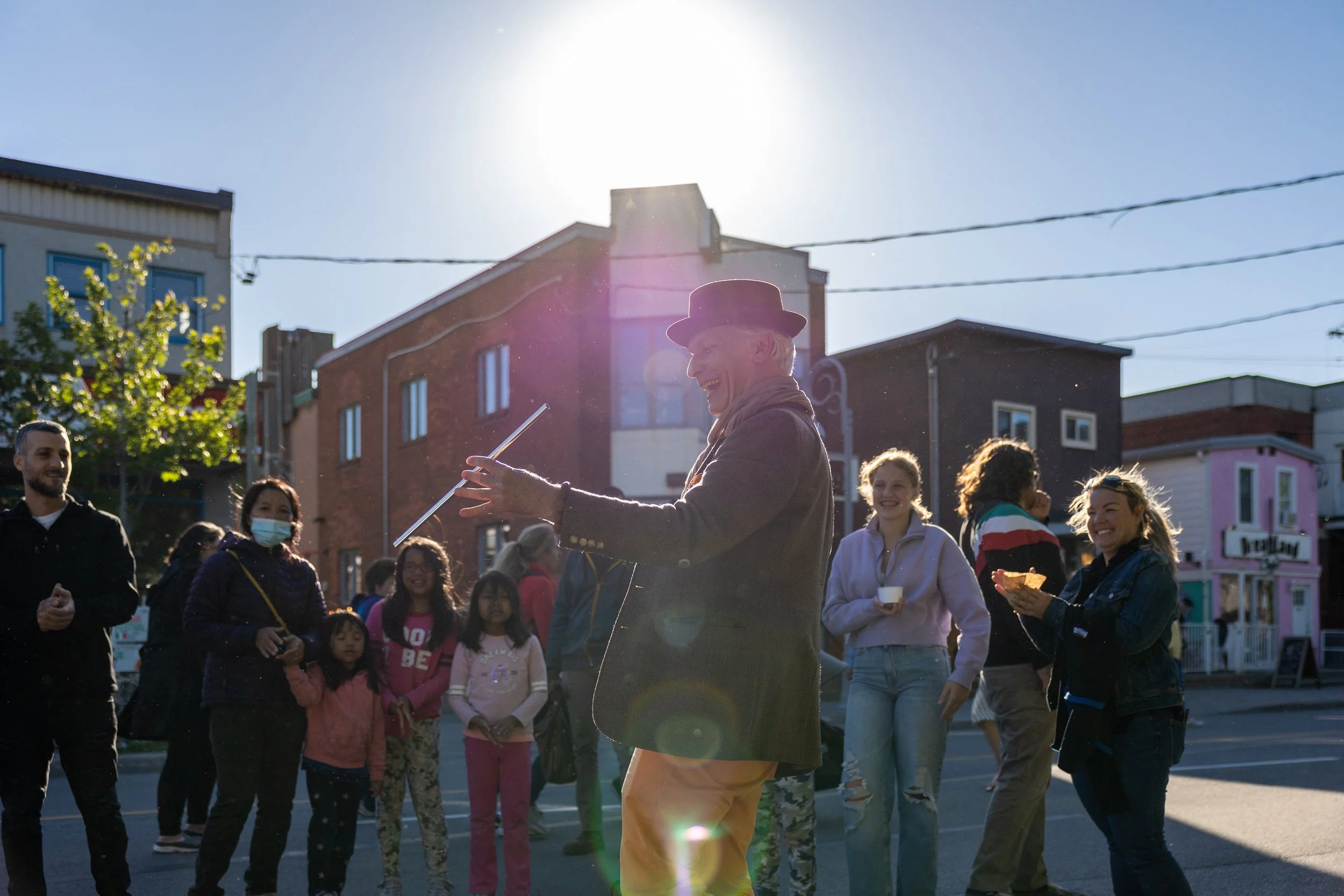 A street performer in a hat entertains a crowd in sunlight, with people smiling and clapping.
