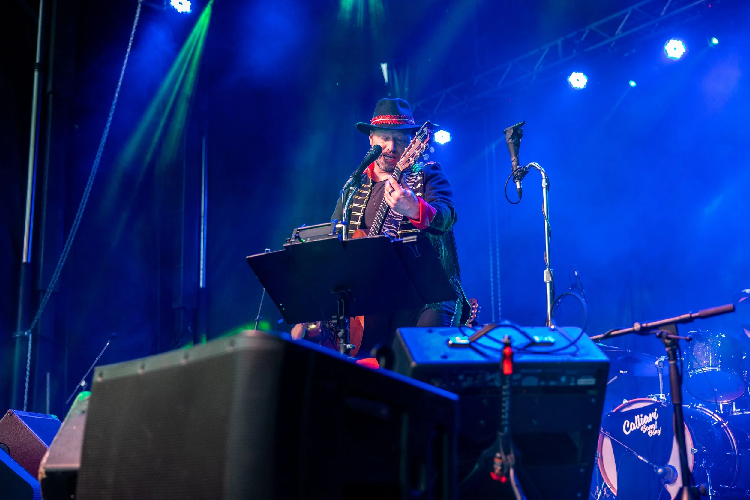 Musician playing guitar on stage with blue lighting and a microphone, wearing a hat and colorful clothing, during a live performance.