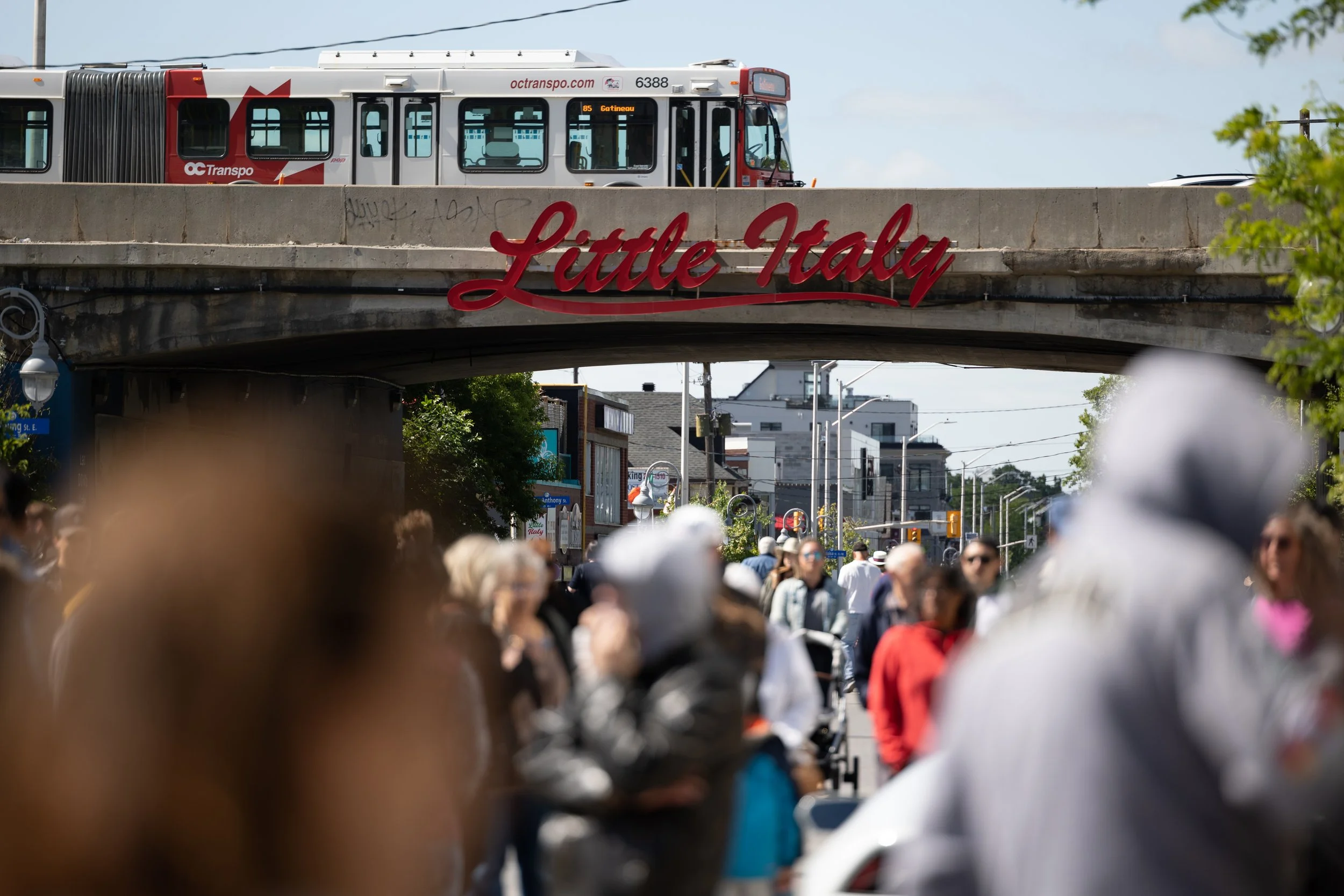 Street scene of Little Italy in Ottawa, Canada, with a crowded pedestrian pathway under a bridge carrying an OC Transpo bus.
