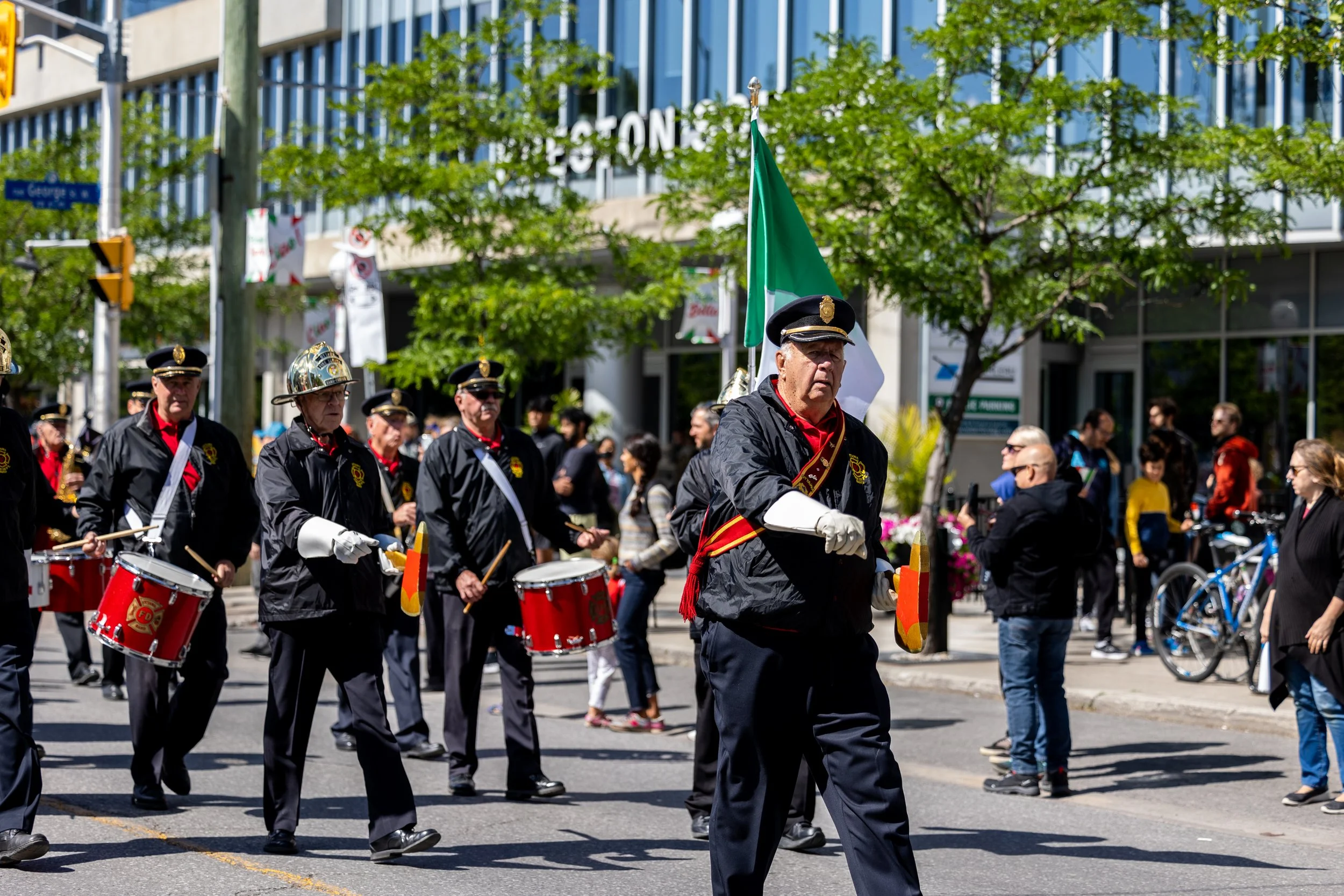 Ottawa Fire Services Band marching in uniform, playing drums and carrying flags, on Preston Street with spectators.