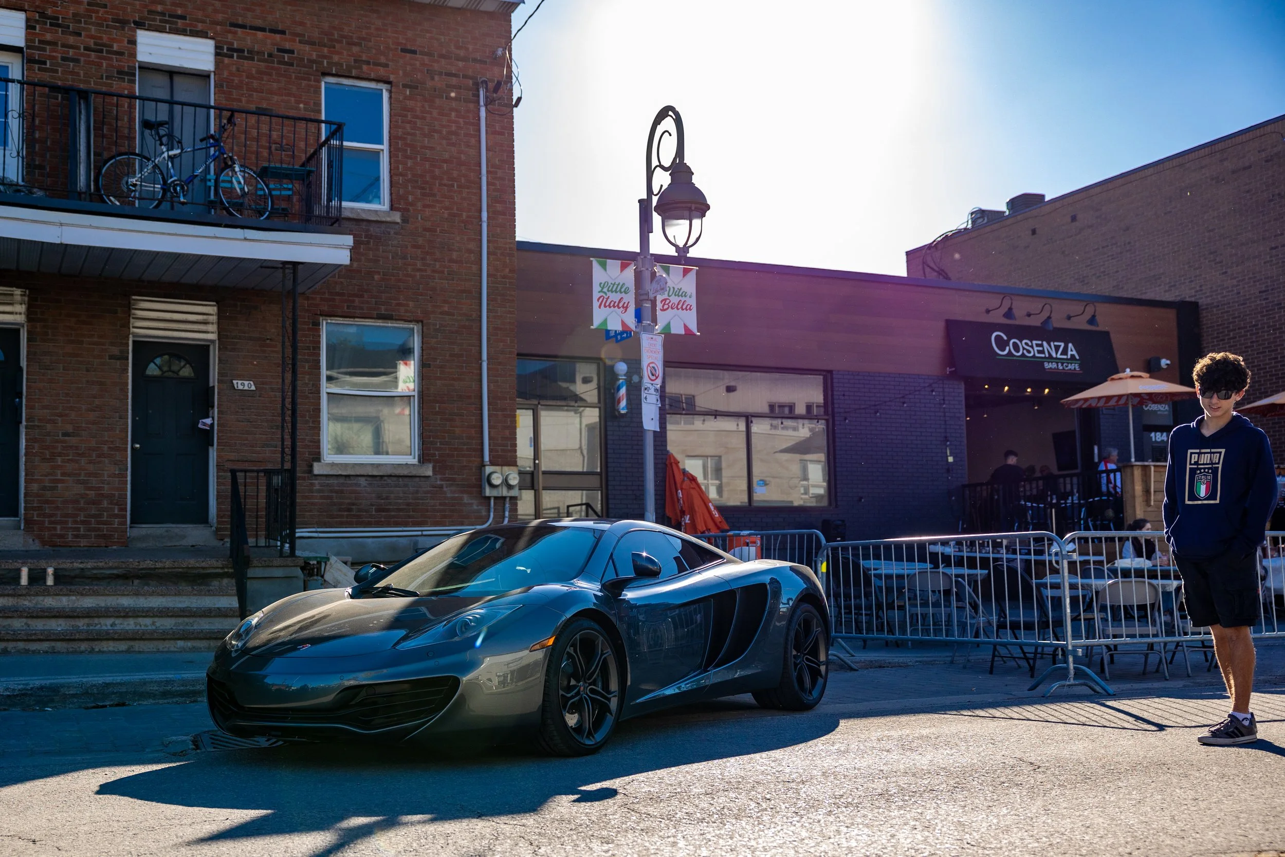 Luxury sports car parked on a street in front of a brick building and a cafe with outdoor seating, under a clear blue sky.