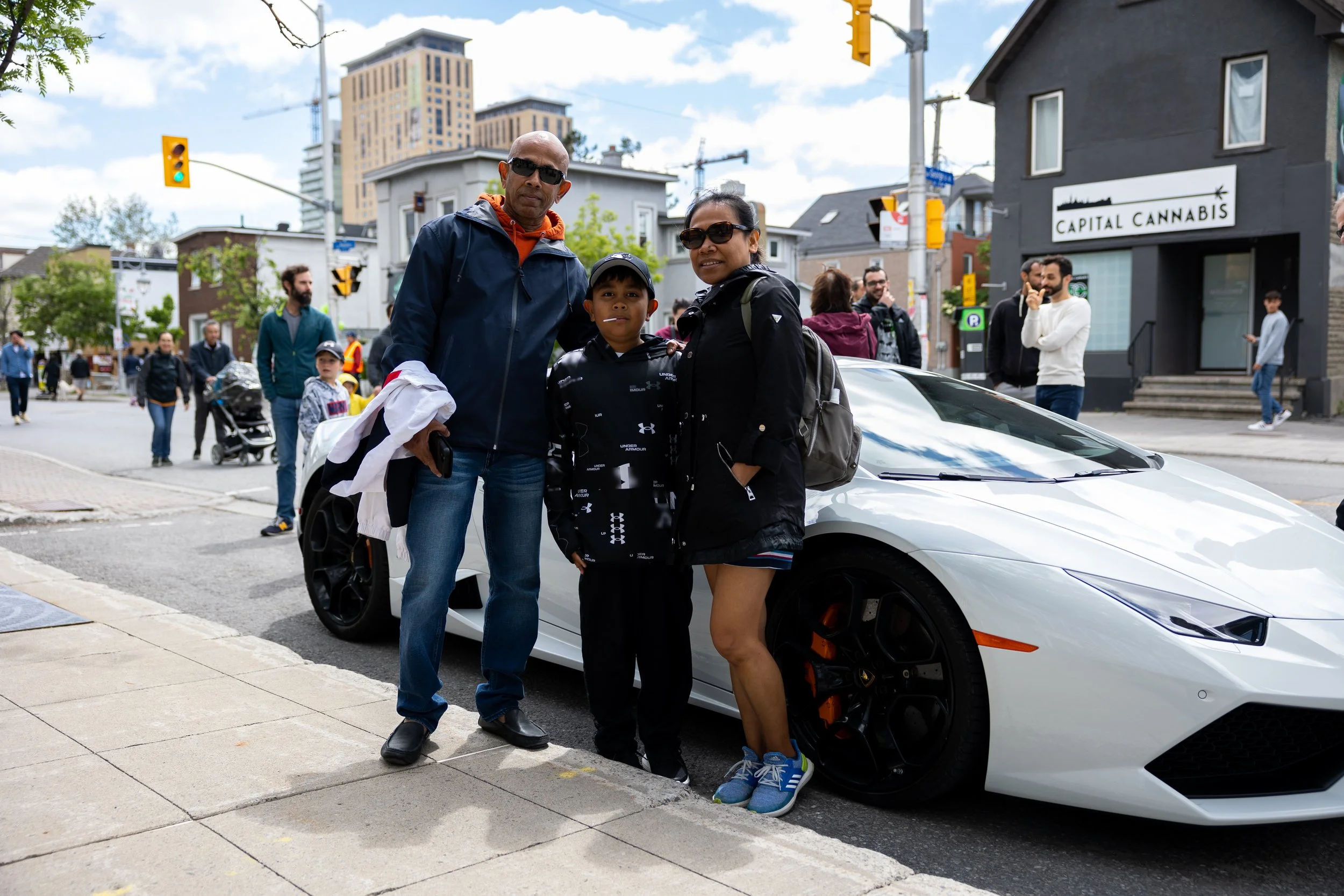 A family of three posing in front of a white sports car on a busy street, with people walking by and buildings in the background.