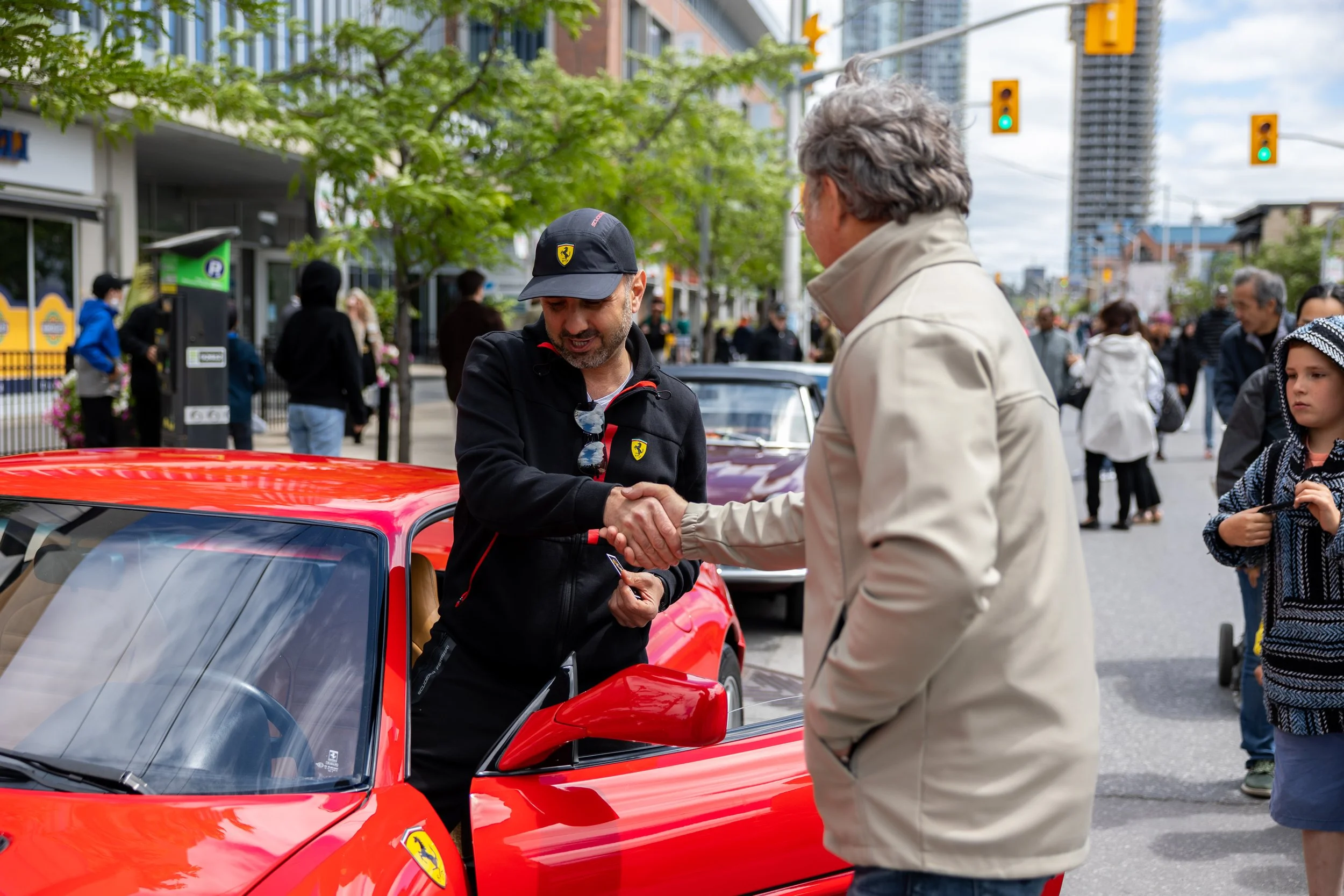 Two men shaking hands beside a red Ferrari car on a busy street with pedestrians in the background.