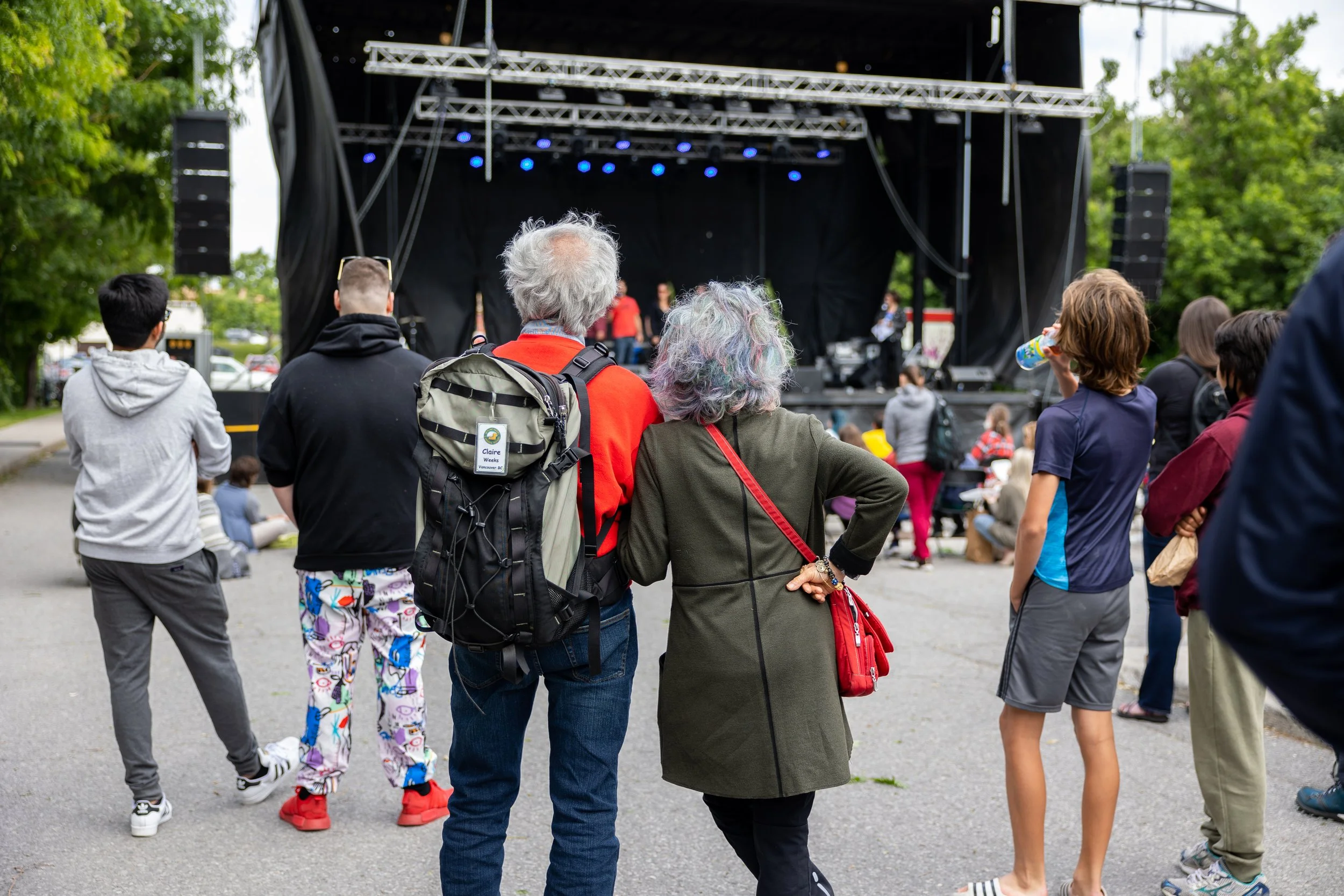 A group of people standing and watching a live outdoor concert on a stage, with speakers and lights overhead.