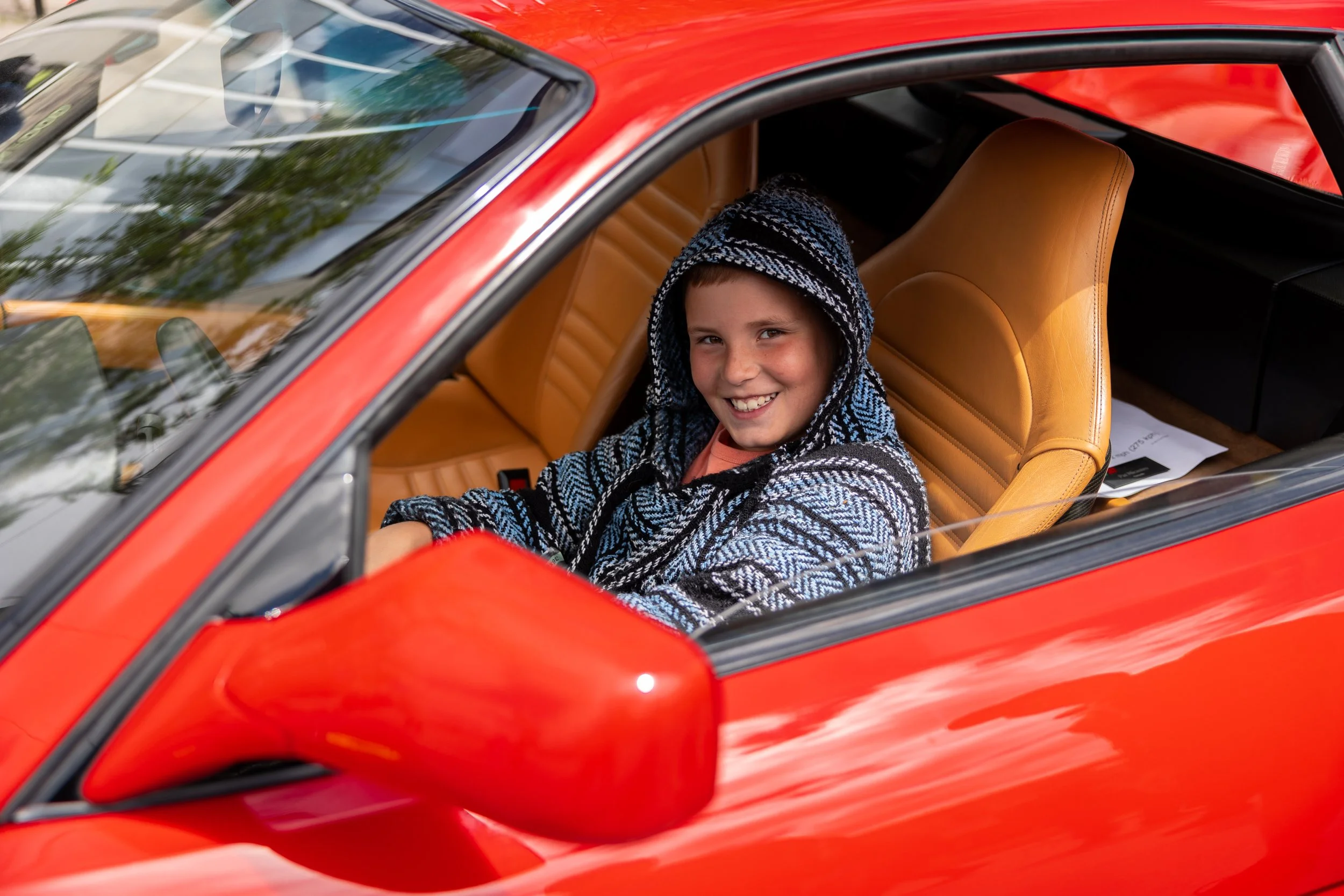 Child smiling in a red sports car with a hooded sweater inside.