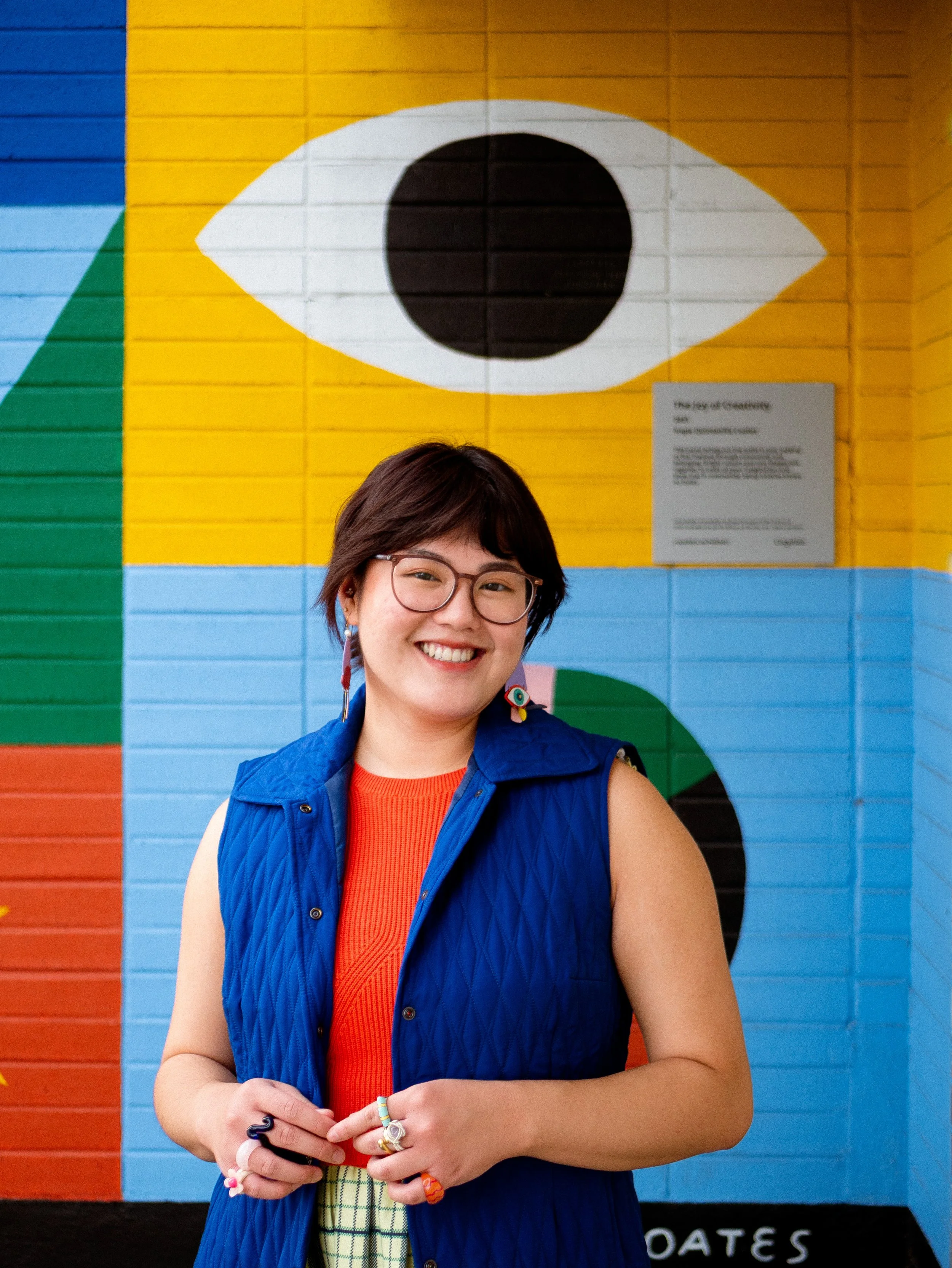 A portrait of Fyonna smiling in front of a colorful mural that features an eye painted on a brick wall.