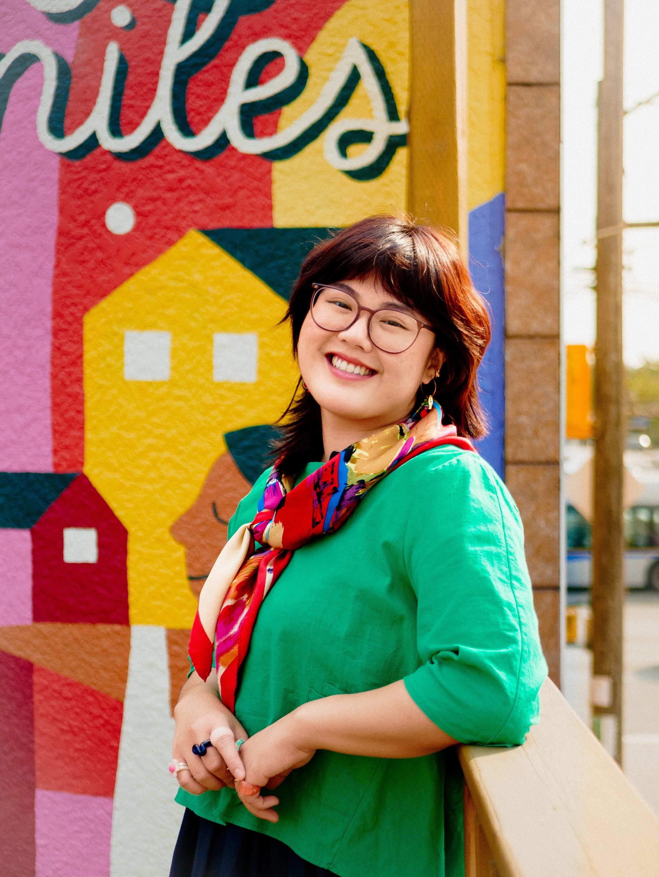 A portrait of Fyonna smiling and leaning against a wooden handrail in front of her City Smiles mural.