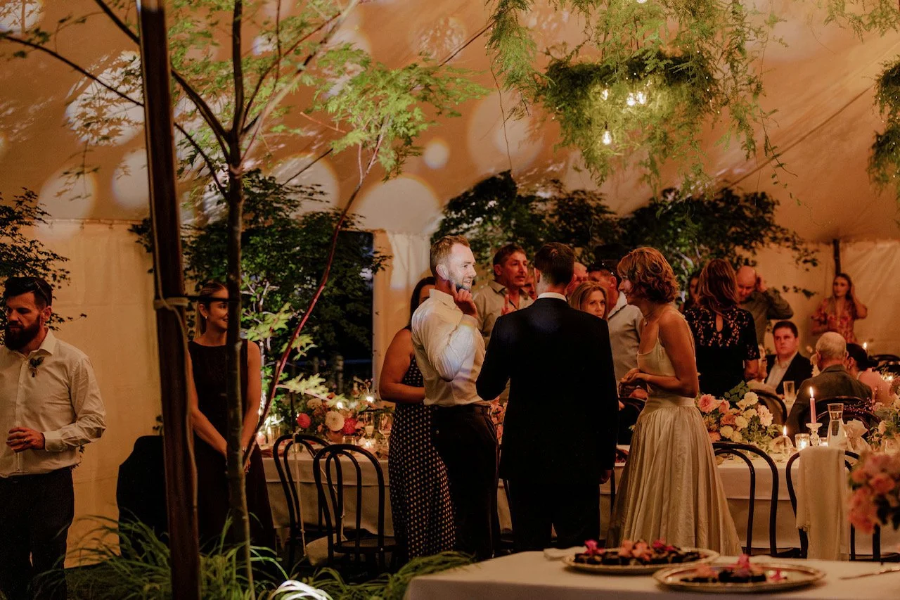 People socializing at a formal event or wedding reception inside a decorated tent with green plants and warm lighting.