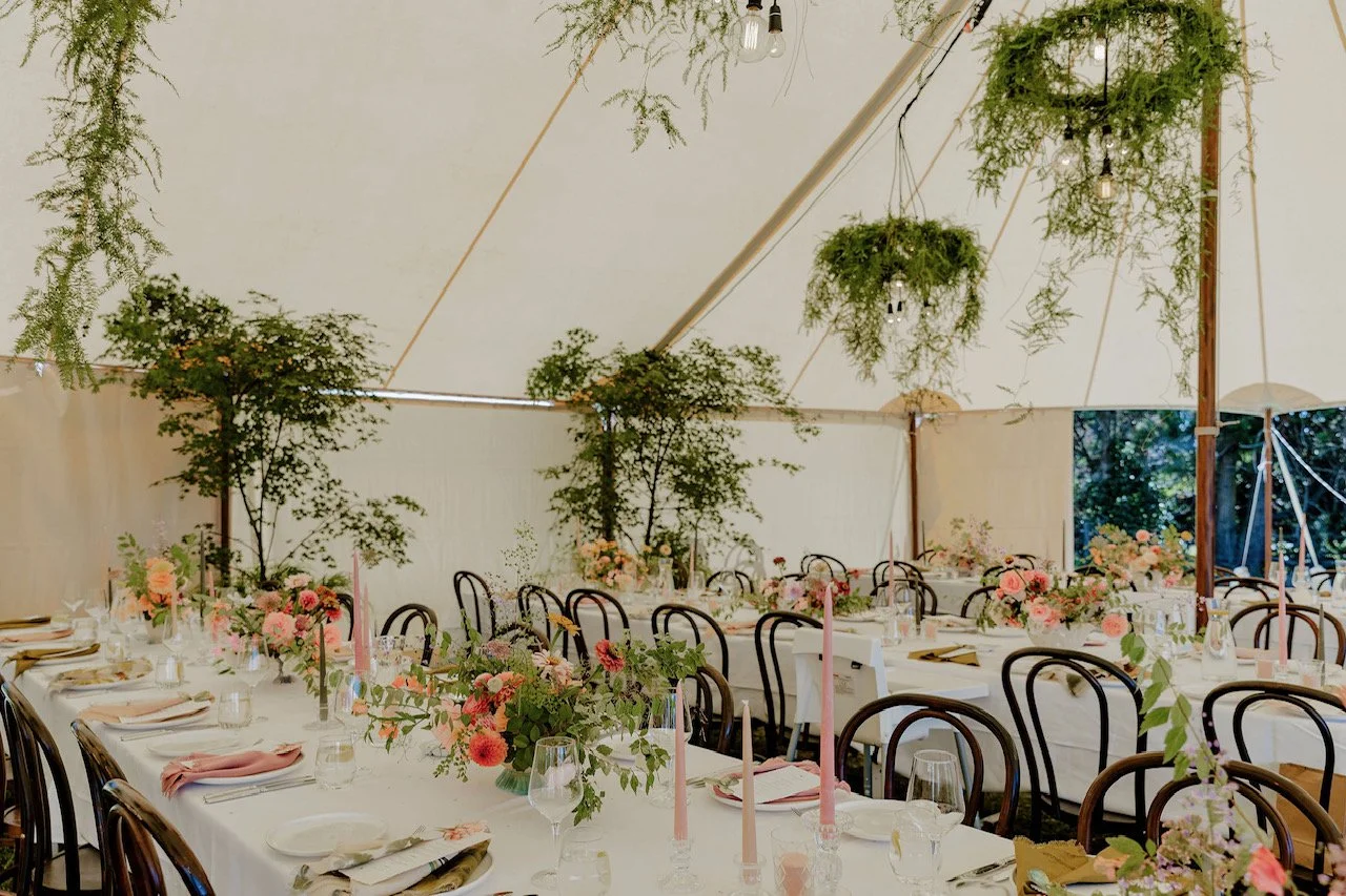 A decorated wedding or event reception tent with long tables adorned with pink and peach flower arrangements, tall pink candles, glasses, plates, and napkins. Hanging greenery and potted plants decorate the tent ceiling, creating a lush, elegant atmo