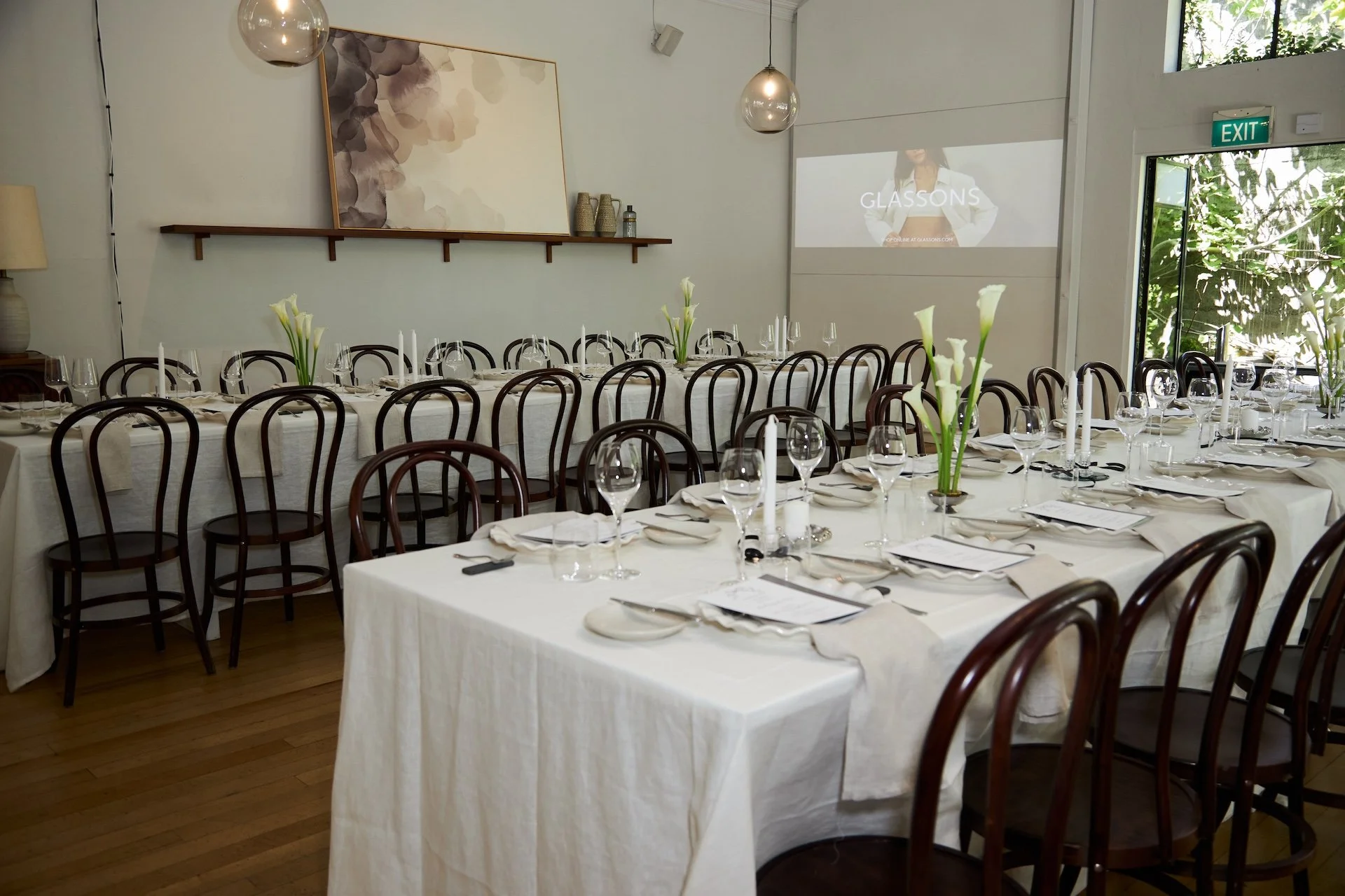 A banquet hall set up for a formal event with multiple long tables covered in white tablecloths, decorated with white calla lily floral centerpieces, wine glasses, and white napkins, with a projector screen displaying the word "GLASSONS" in the background.