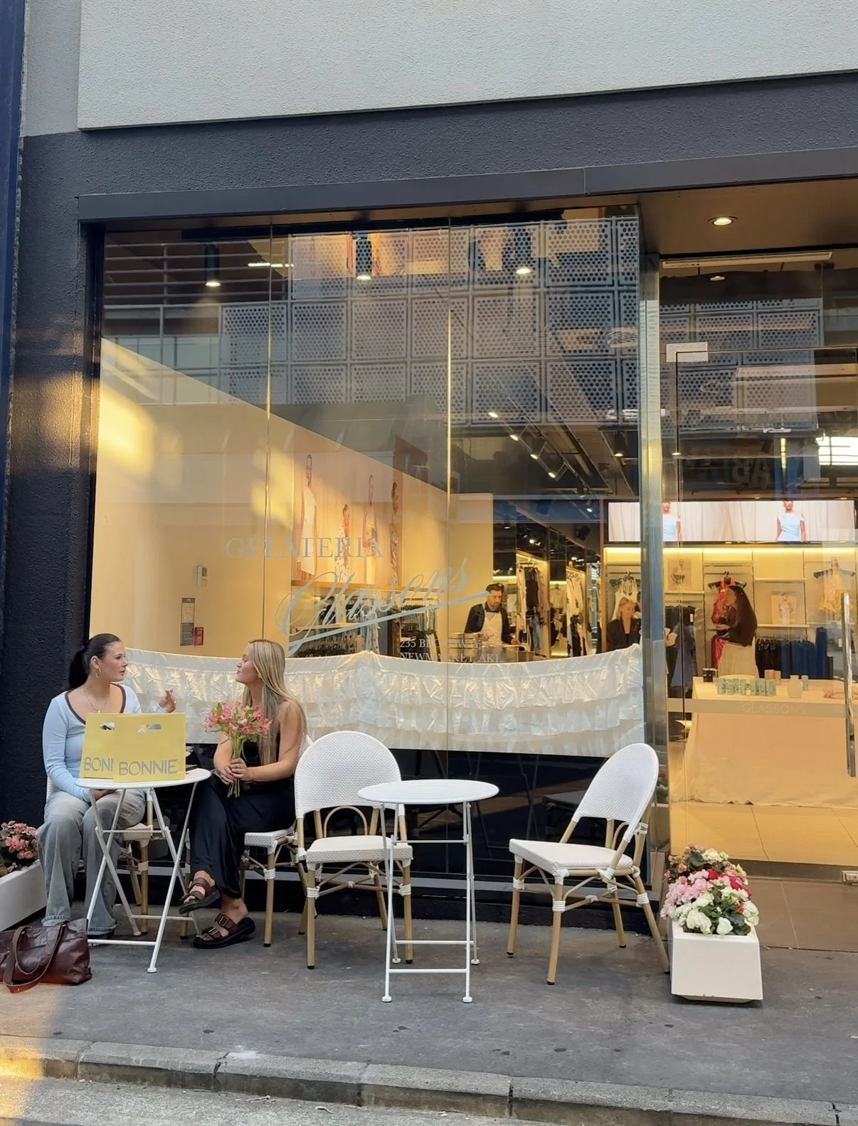 Two women are sitting outside a clothing boutique called 'Galleria Cherby,' conversing. They have pink flowers in a black vase and a yellow shopping bag labeled 'Bon Bon' on a small white table between them. The storefront features large glass windows with mannequins and clothing inside, and there are floral planters on each side of the sidewalk.