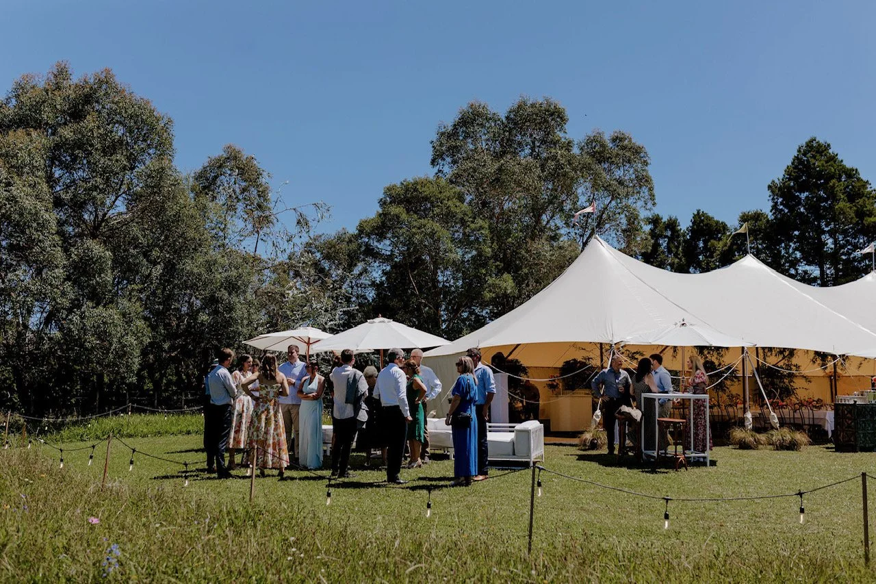 People gathered outside under large white tents and umbrellas at an outdoor event on a sunny day, with trees and blue sky in the background.