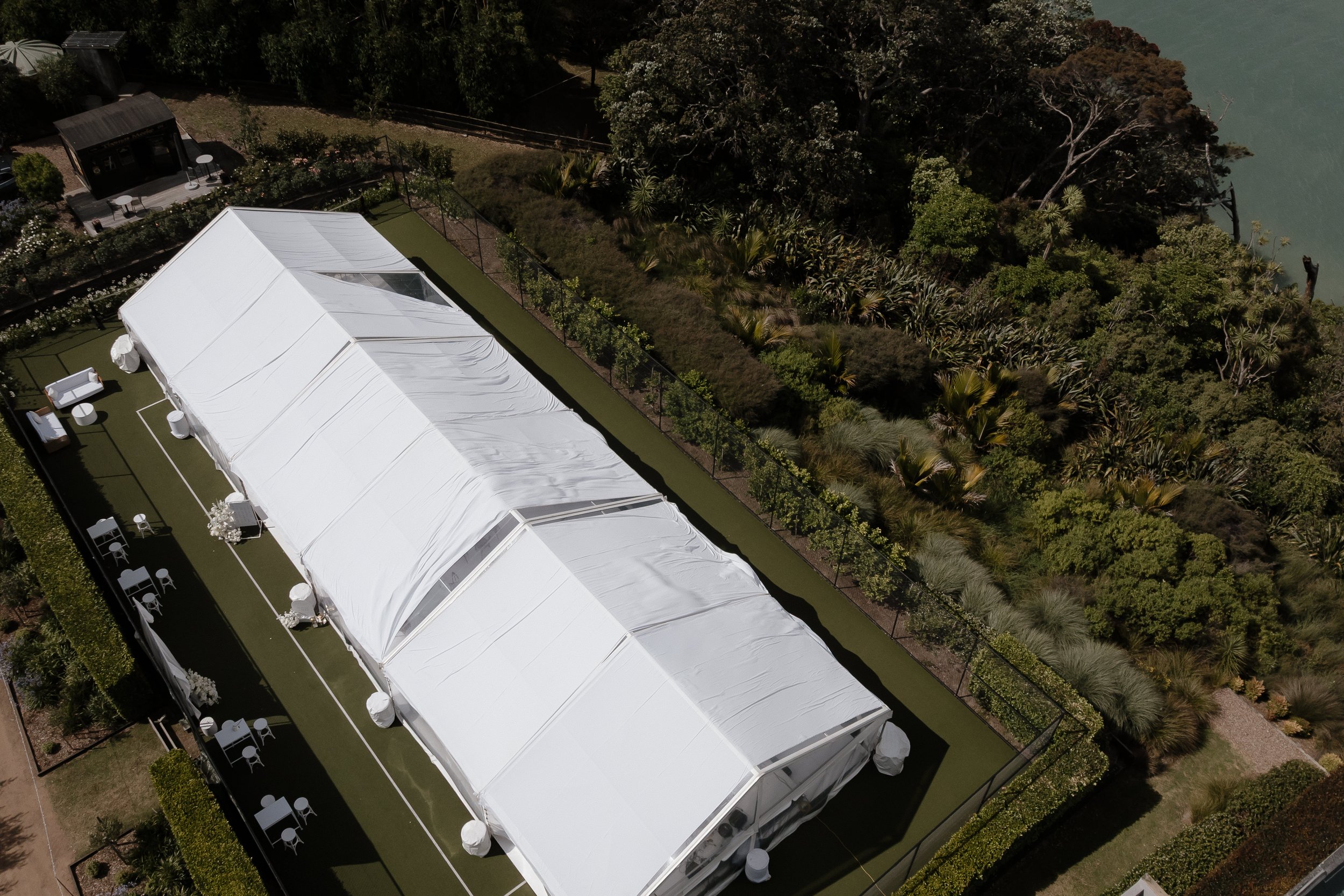 Aerial view of a large white event tent set up on a grassy area next to a lush garden with trees and shrubs, adjacent to a body of water.