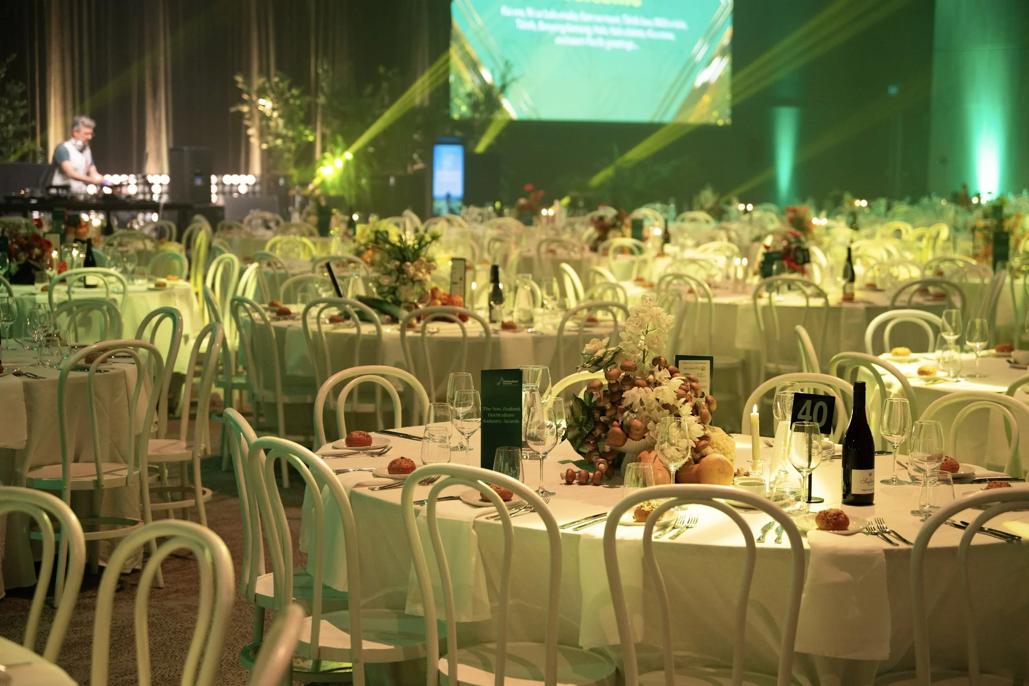 A banquet hall set up with round tables decorated with floral centerpieces, wine bottles, and place settings, ready for an event. Stage with a large screen and DJ playing music is visible in the background, with colorful lighting effects.