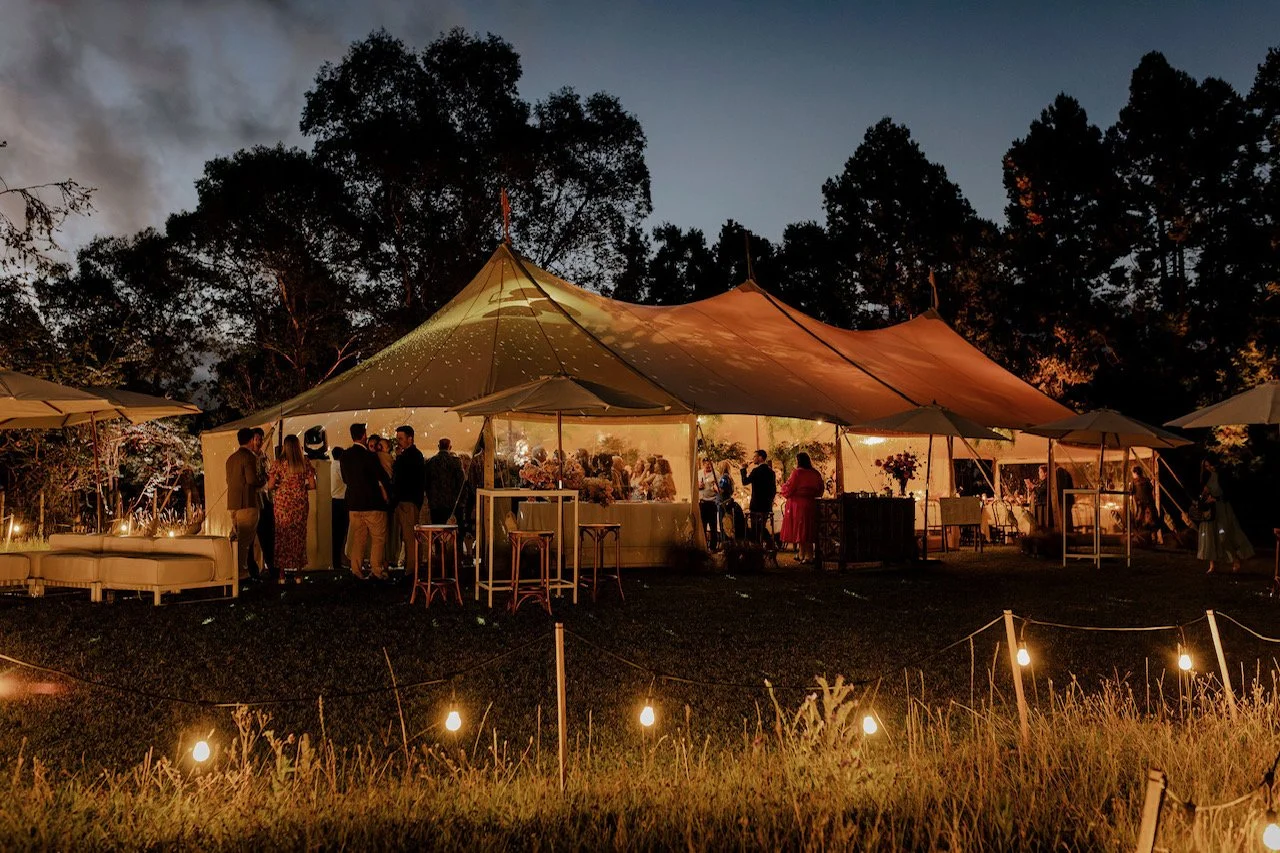 An outdoor evening event under a large tent with people socializing, decorated with string lights and surrounded by trees.