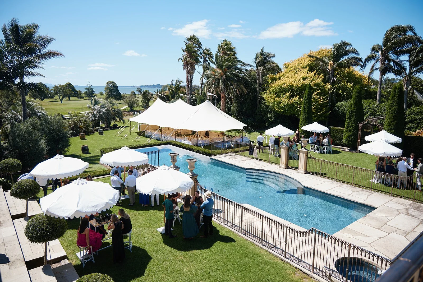 Guests gathered around pools and under white umbrellas at an outdoor event on a sunny day, with lush greenery and trees in the background.
