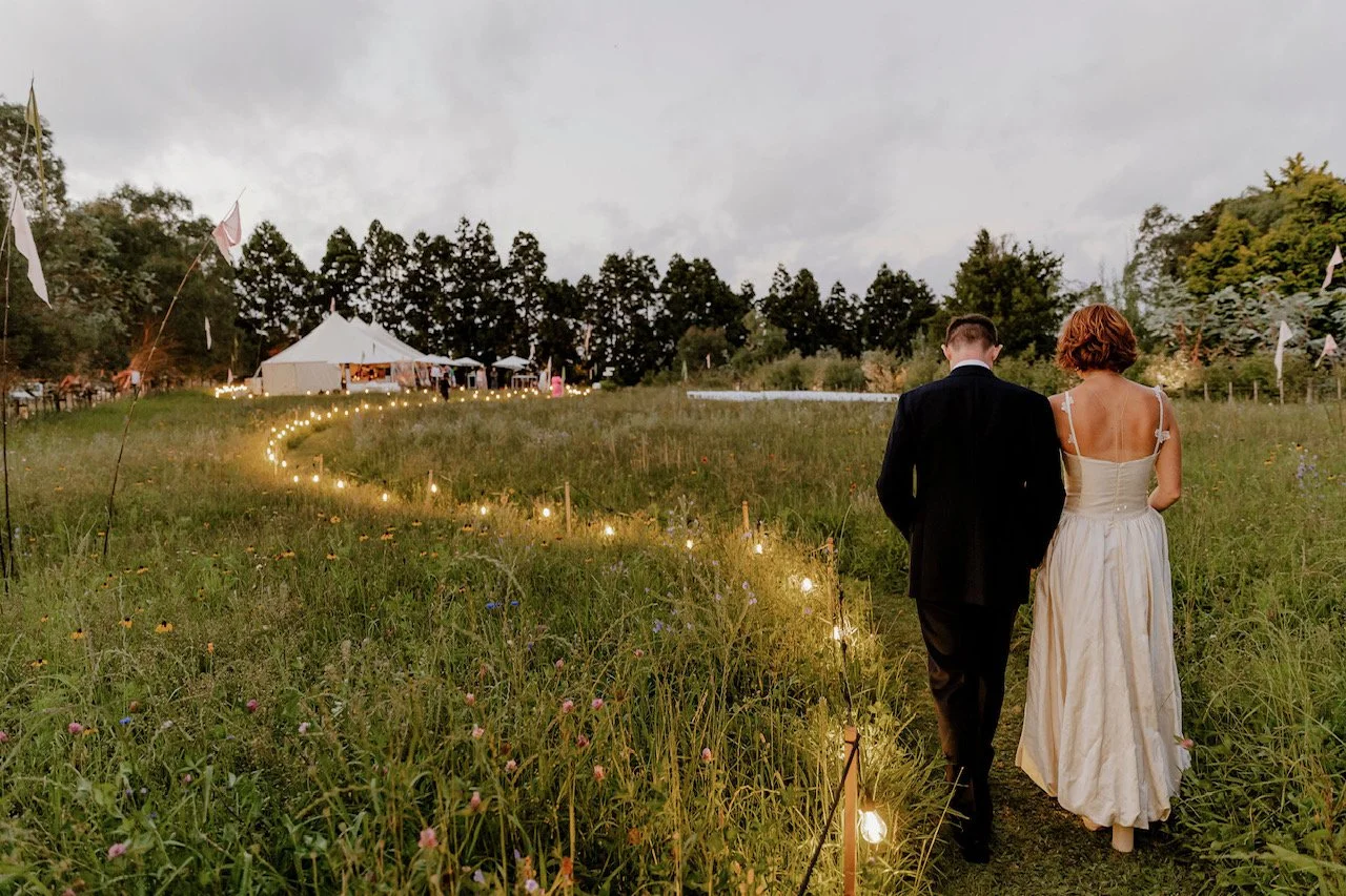 A bride and groom walking along a lit pathway in an open field at sunset, leading to a wedding reception tent in the background.