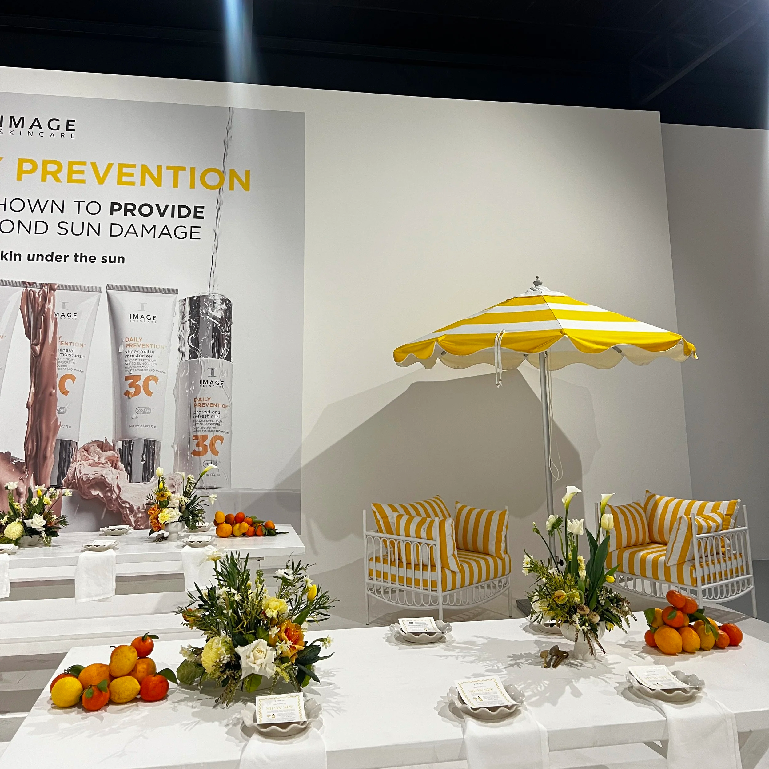 A decorated dining display with a sunny yellow and white striped umbrella, matching striped chairs, floral arrangements, and an assortment of citrus fruits on a white table in front of a large skincare advertisement.