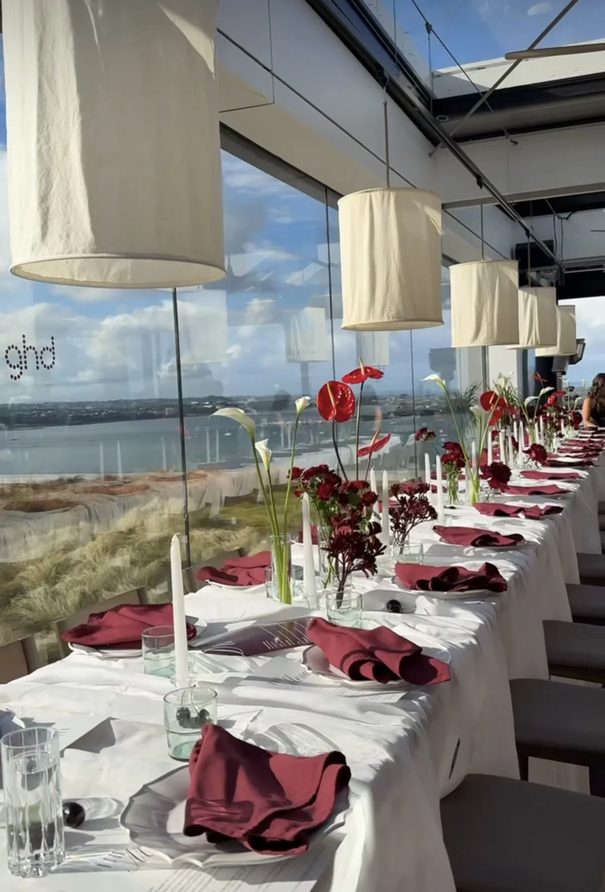 Elegant dining table set for a meal with white tablecloths, pink napkins, candles, and floral centerpieces. Large windows with a view of the water and sky, hanging lamps above.