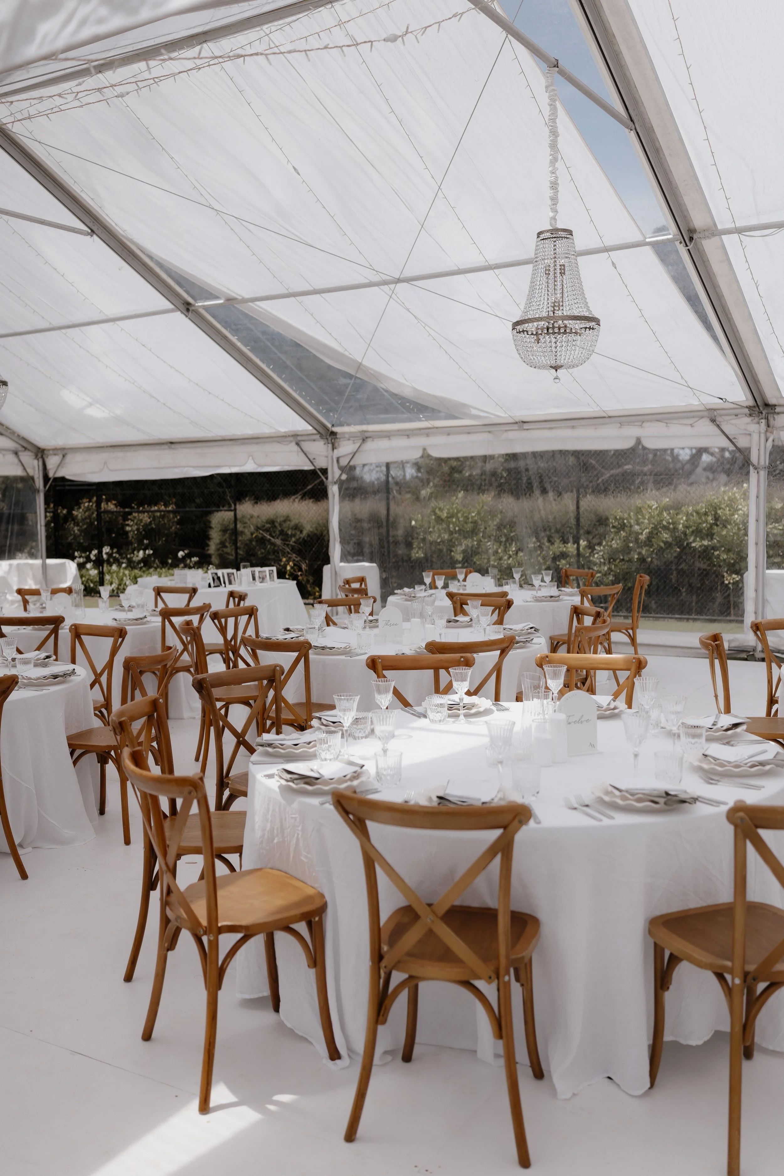 A wedding reception setup inside a white tent with round tables covered in white tablecloths, decorated with glassware and cutlery, surrounded by wooden chairs, and a chandelier hanging from the ceiling.