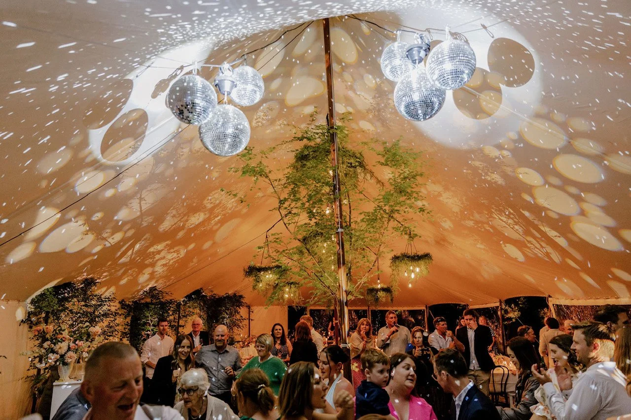 People dancing and socializing inside a large decorated tent with hanging disco balls and warm lighting.