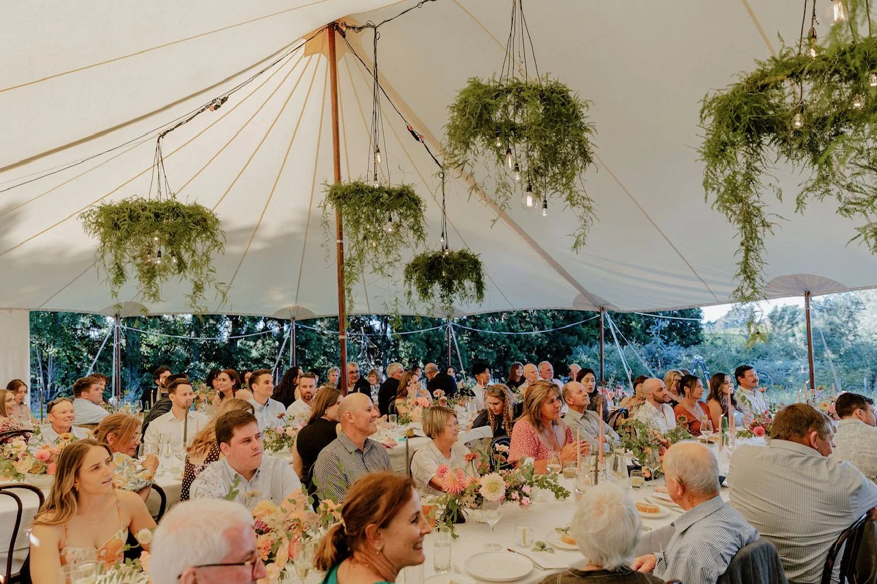 Guests seated at tables under a large tent with hanging greenery and string lights, attending a wedding or celebration event.