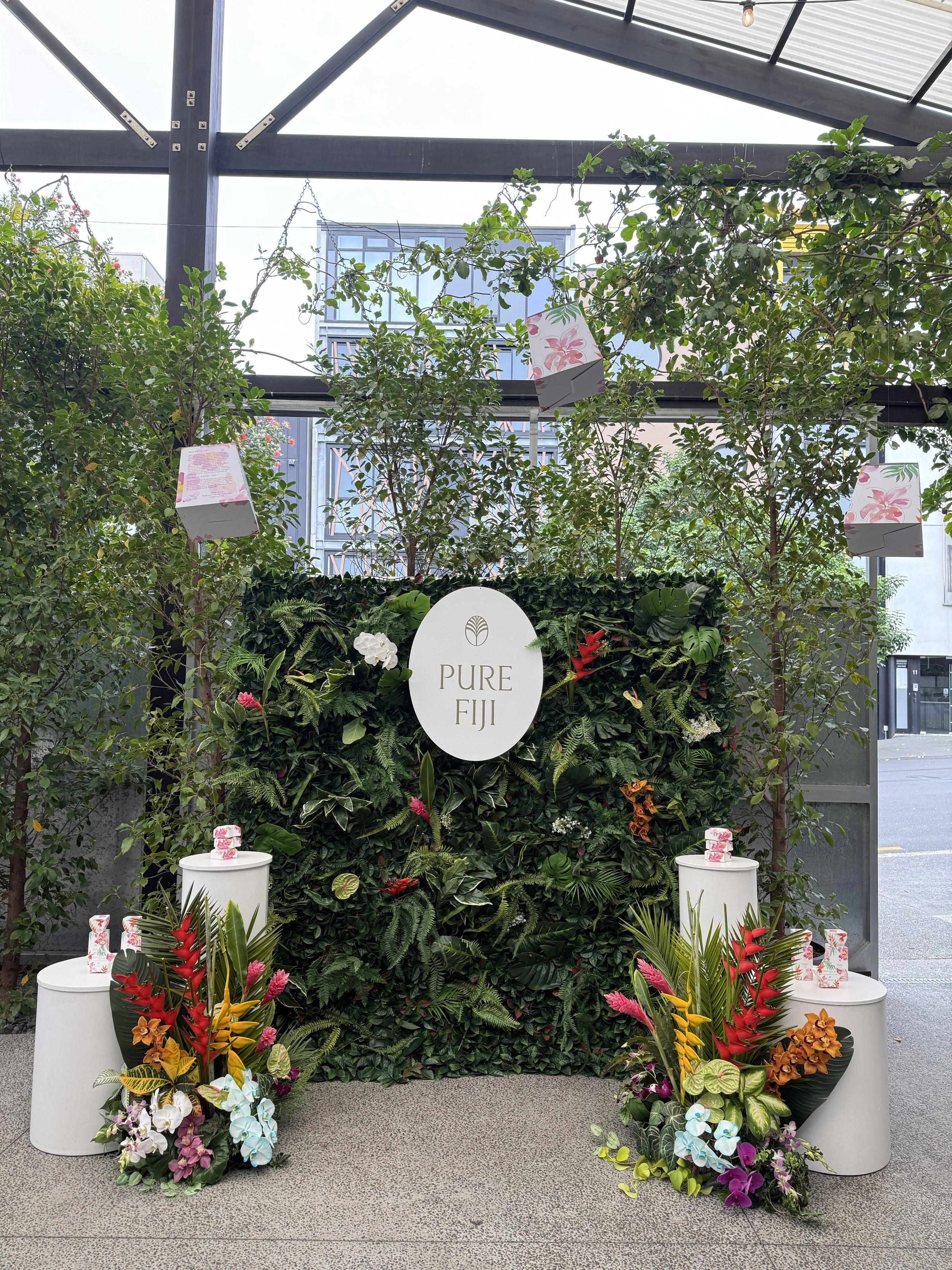 A decorative display at a venue with a lush green plant backdrop, a white 'Pure Fiji' logo sign, colorful tropical flowers, and white cylindrical pedestals with floral arrangements.