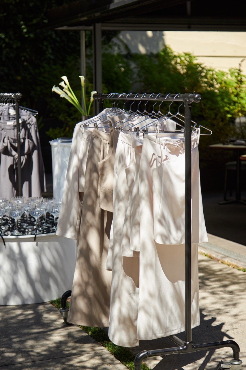 Outdoor clothing rack with beige pants and skirts, alongside table with glassware and potted calla lilies.