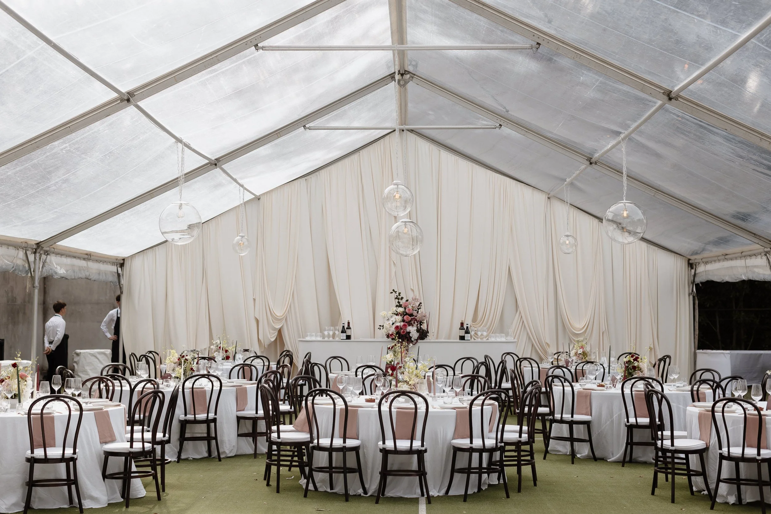 Elegant event tent with round tables decorated with white tablecloths and blush pink runners, set with wine glasses, silverware, and floral centerpieces in pink, cream, and burgundy. Light fixtures hang from the ceiling, and white drapes adorn the walls. There are a few people in formal attire standing inside the tent, indicating a celebration or wedding reception.