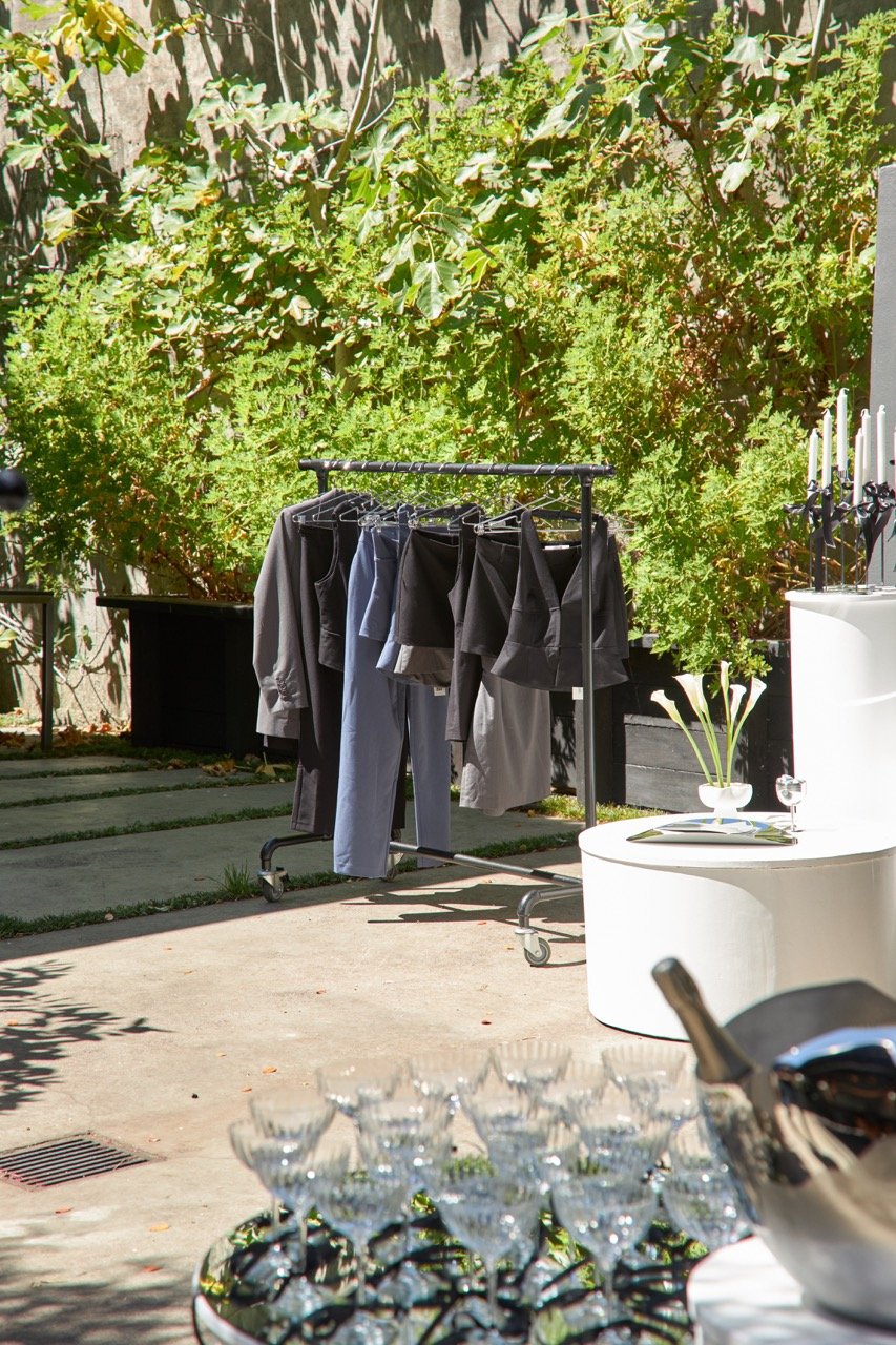 Outdoor scene with a clothing rack displaying black and gray garments, surrounded by greenery. In the foreground, a table holds multiple empty glasses and a silver ice bucket.