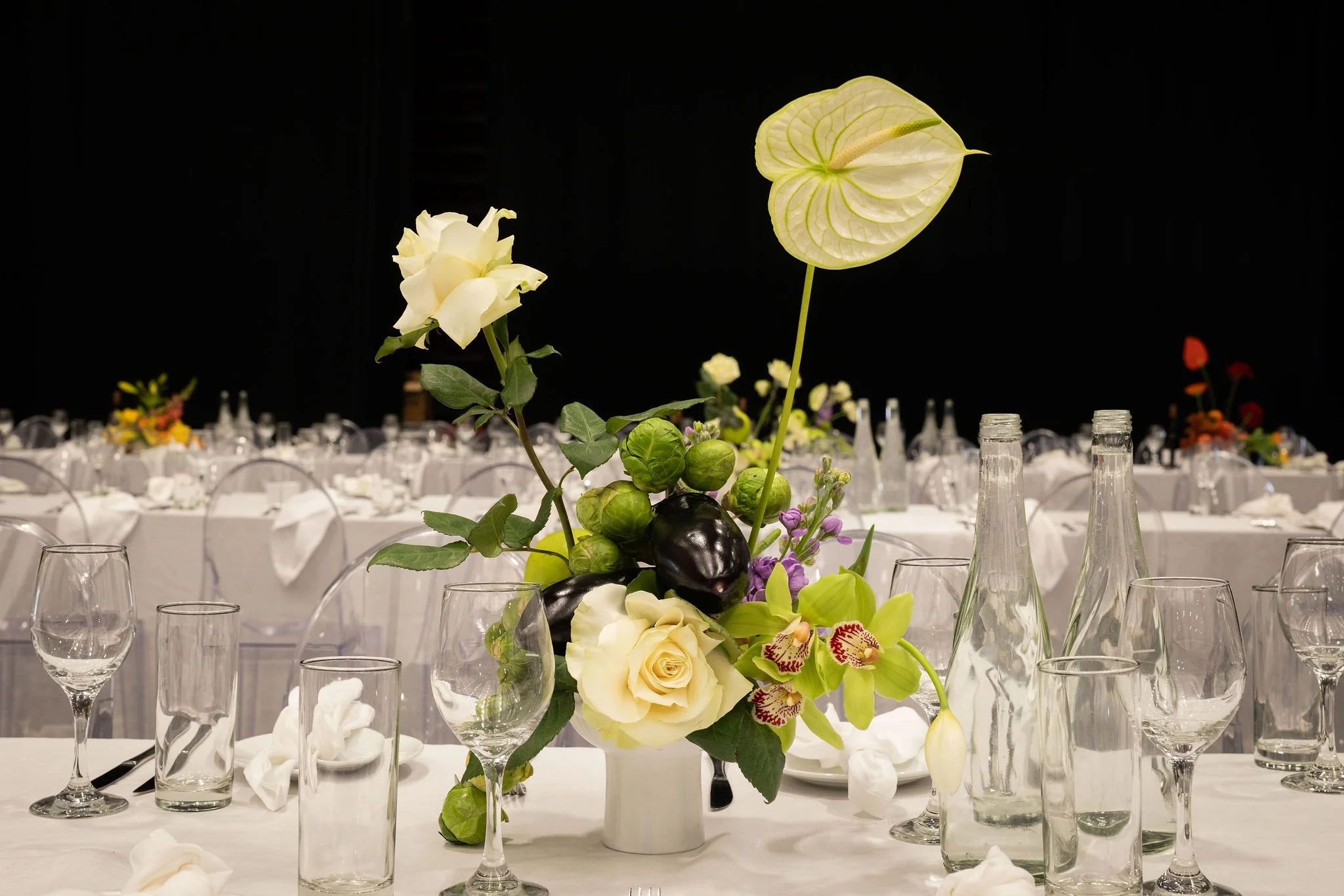 Elegant table centerpiece with white roses, anthurium, and green leaves on a white tablecloth surrounded by empty glassware at a formal event.