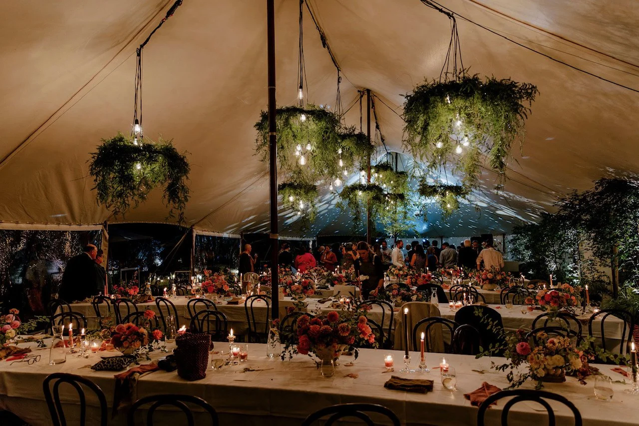 A decorated event tent with hanging greenery and light bulbs, long tables with floral centerpieces, candles, and guests in the background.