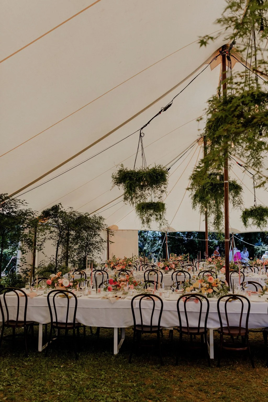 A decorated outdoor dining area under a large white tent with hanging plants, floral centerpieces, and black chairs around long tables.