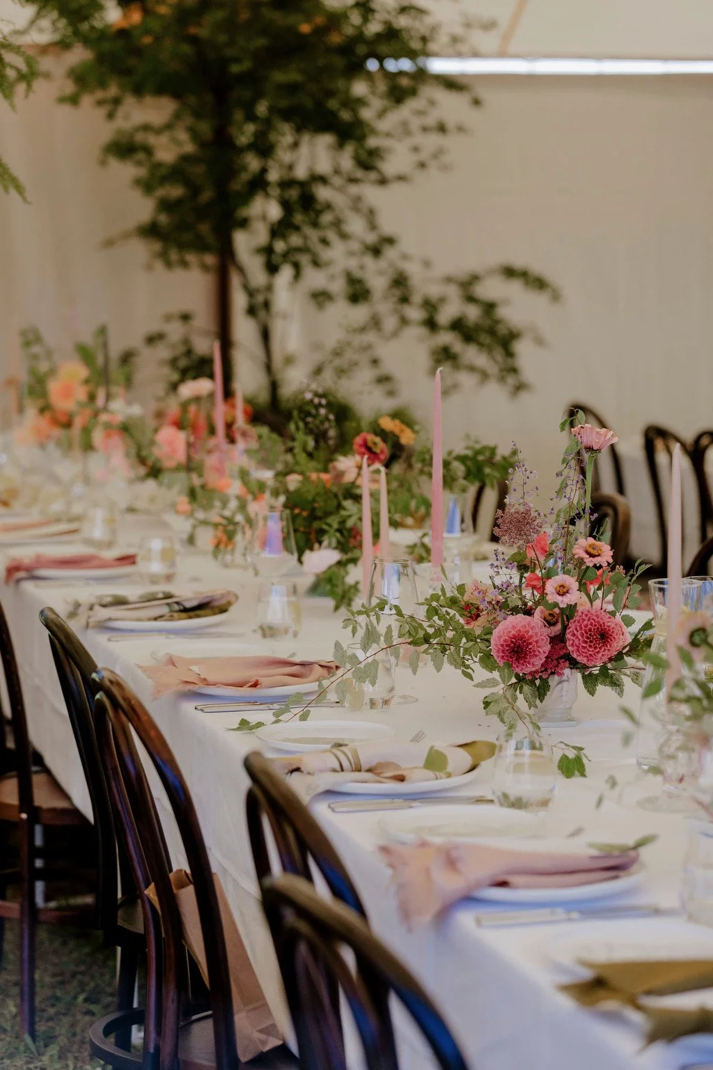 A long dining table decorated with pink and peach flowers, tall pink candles, and place settings with napkins and glasses, set up for an event in a room with a plant and chairs in the background.