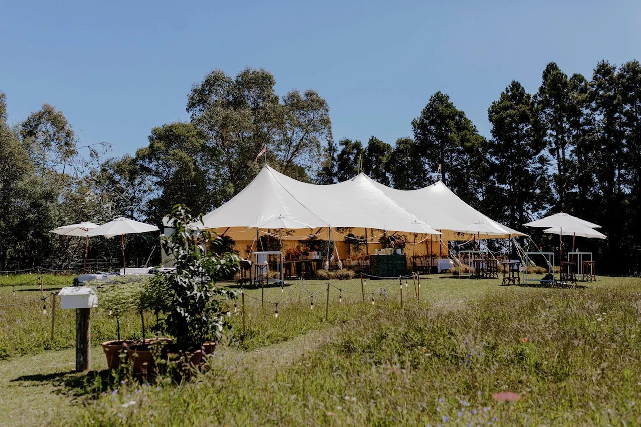 Large white event tent with tables and chairs underneath, surrounded by smaller umbrellas and outdoor seating, set on a grassy field with trees in the background on a sunny day.