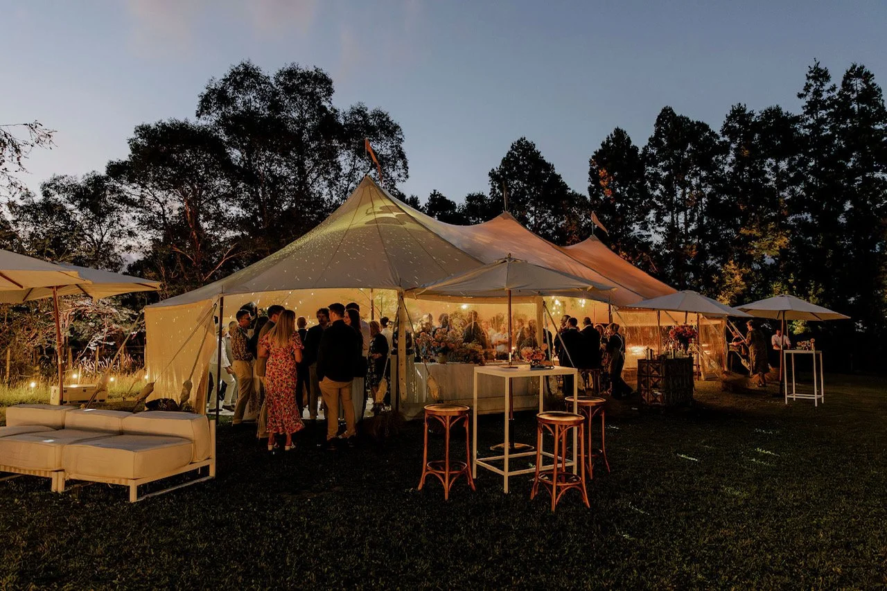 Outdoor evening event with a large white event tent, string lights, people mingling, and outdoor furniture, surrounded by trees.