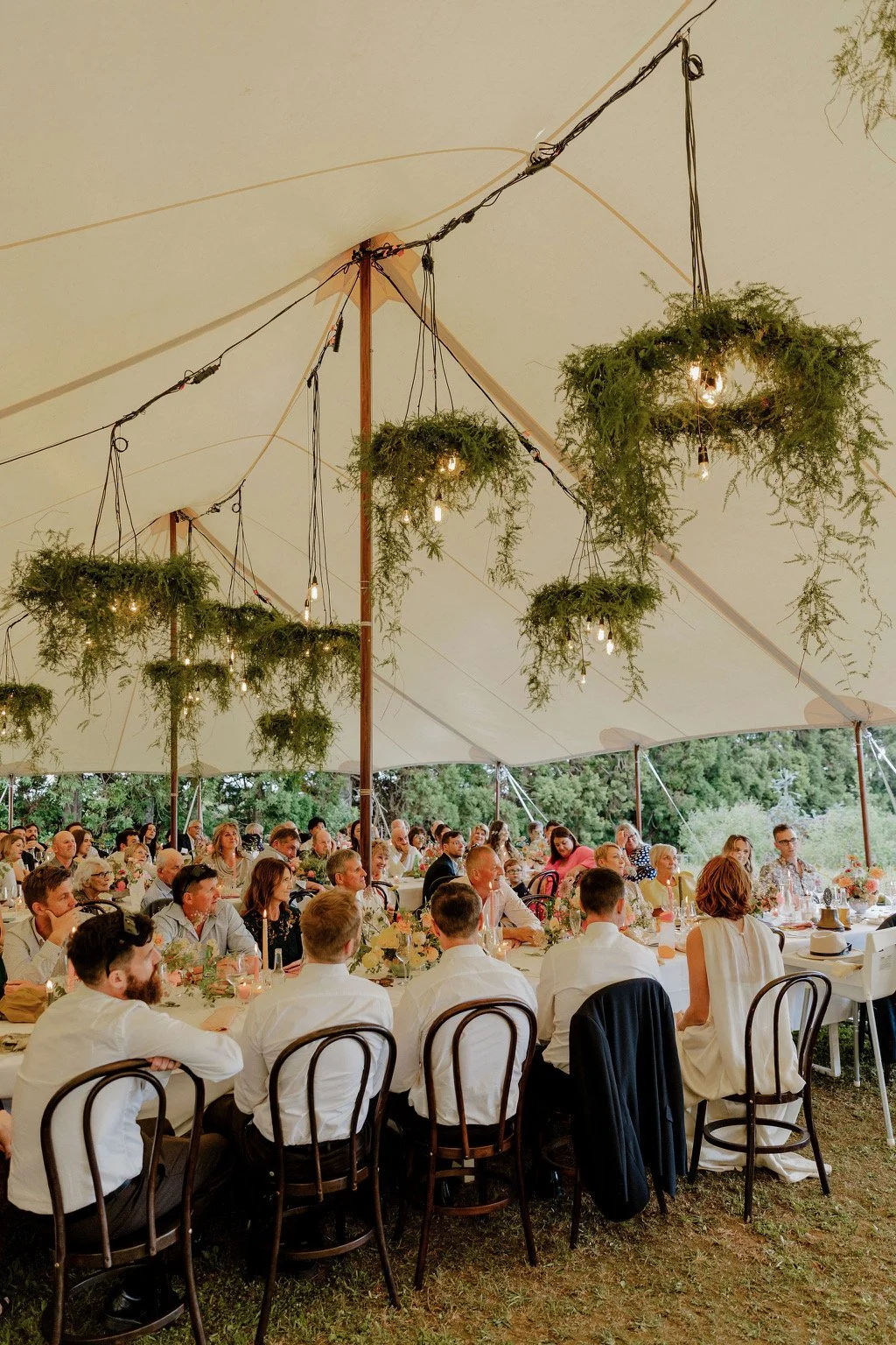People seated at long tables under a large white tent with hanging greenery and lights, attending a gathering or celebration outdoors near trees.