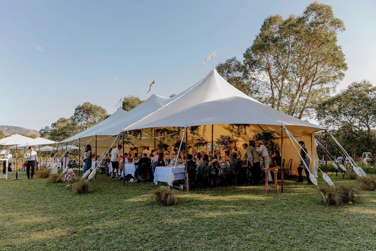 Outdoor gathering under large white tent with people seated at tables, surrounded by trees and greenery.