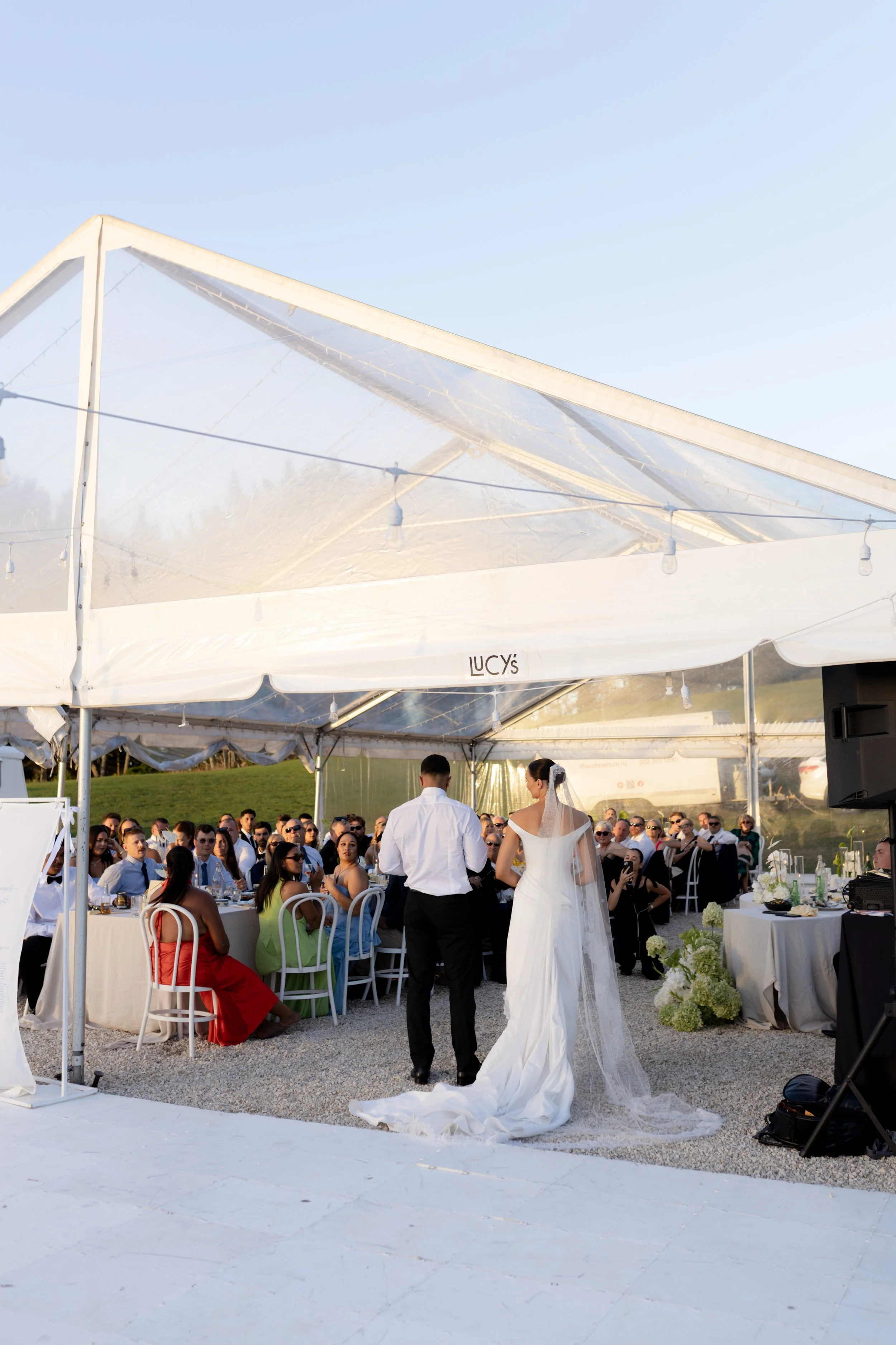 A couple, dressed in wedding attire, standing under a tent as they exchange vows during an outdoor wedding ceremony with guests seated around.