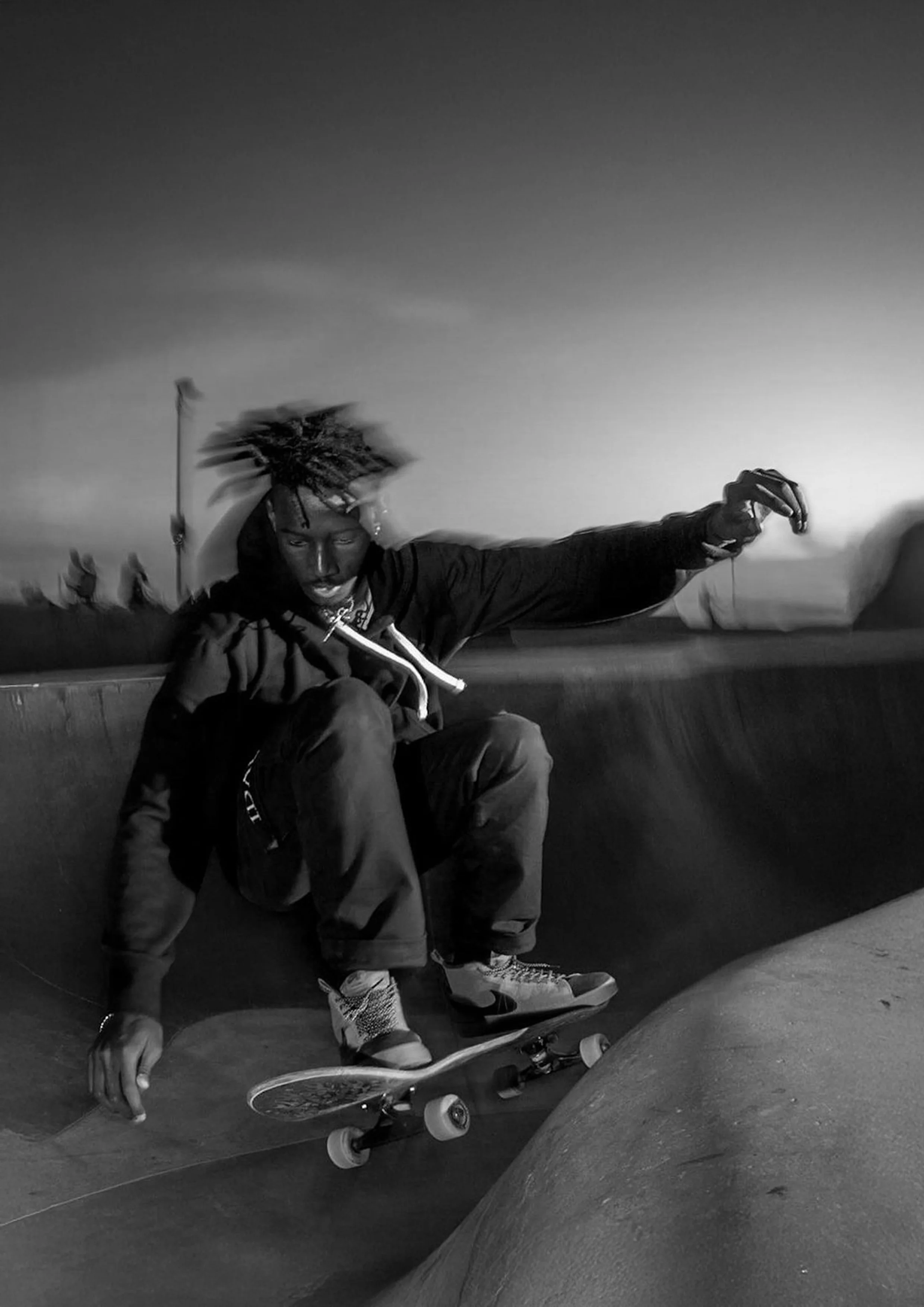 Black and white photo of a skateboarder in mid-trick at a skatepark, with his hair flying and an intense expression.