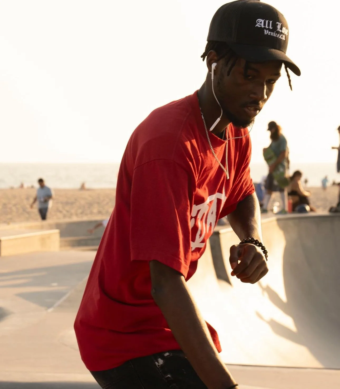 A young man wearing a red t-shirt, black cap, and wristbands, listening to earphones at a skatepark on a sunny day, with a beach and people in the background.