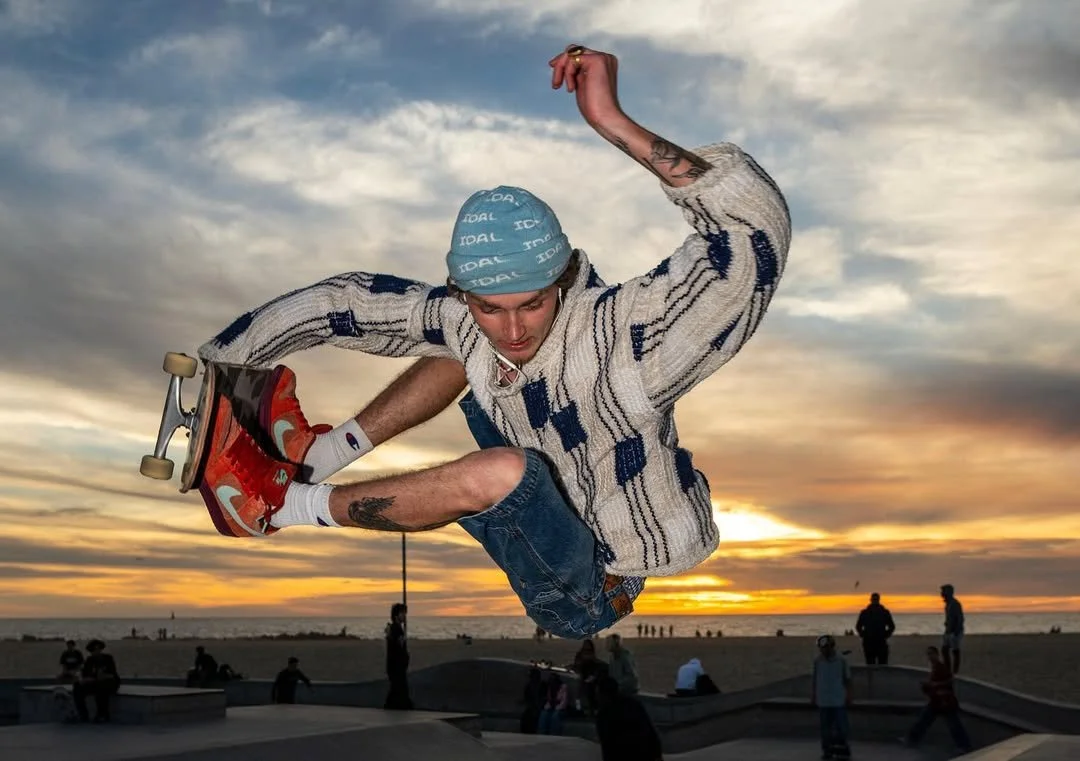 Skateboarder performing a mid-air trick at sunset on the beach, wearing a blue beanie and striped sweater.