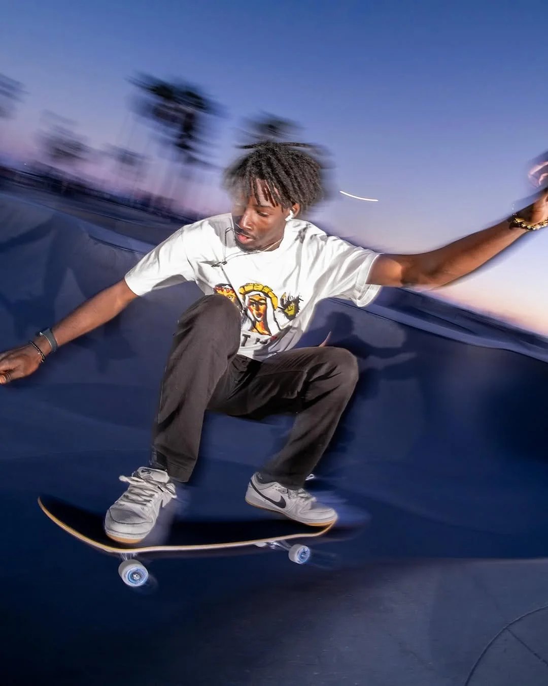 A young man skateboarding in a bowl during dusk, wearing a white graphic T-shirt, gray pants, and white Nike sneakers, with trees and a clear sky in the background.
