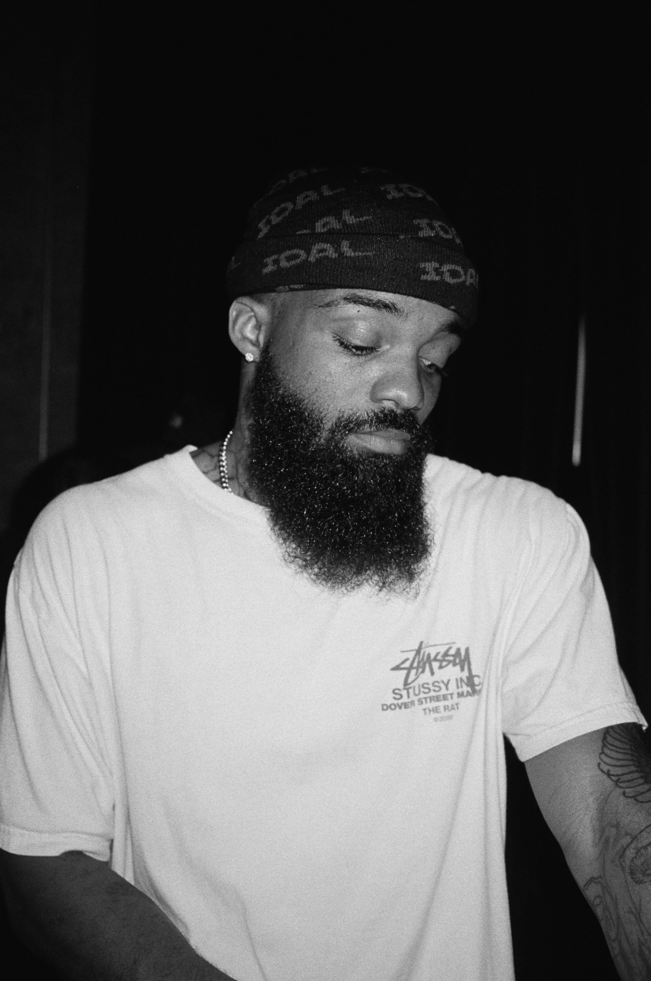 A man with a beard and tattoos wearing a Stussy t-shirt and a beanie hat looking down.