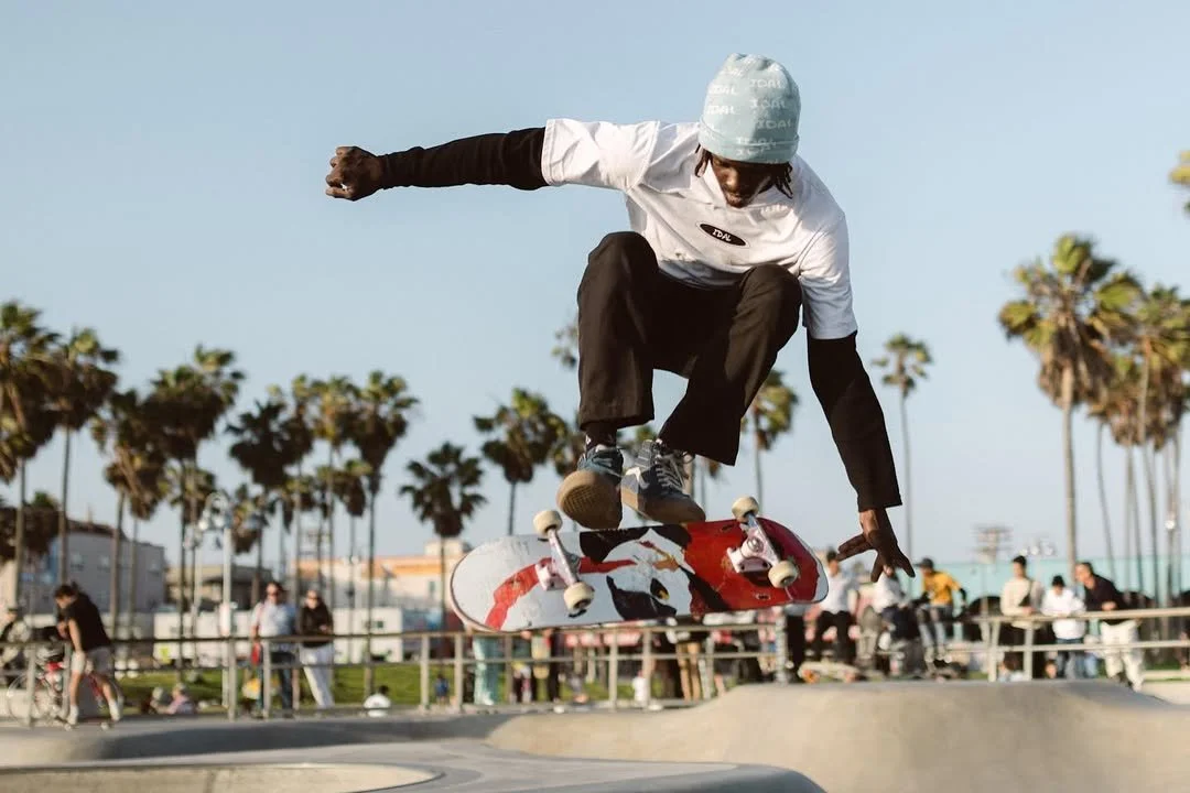 Skateboarder wearing a white helmet and black pants performing a trick at a skate park with palm trees and spectators in the background.