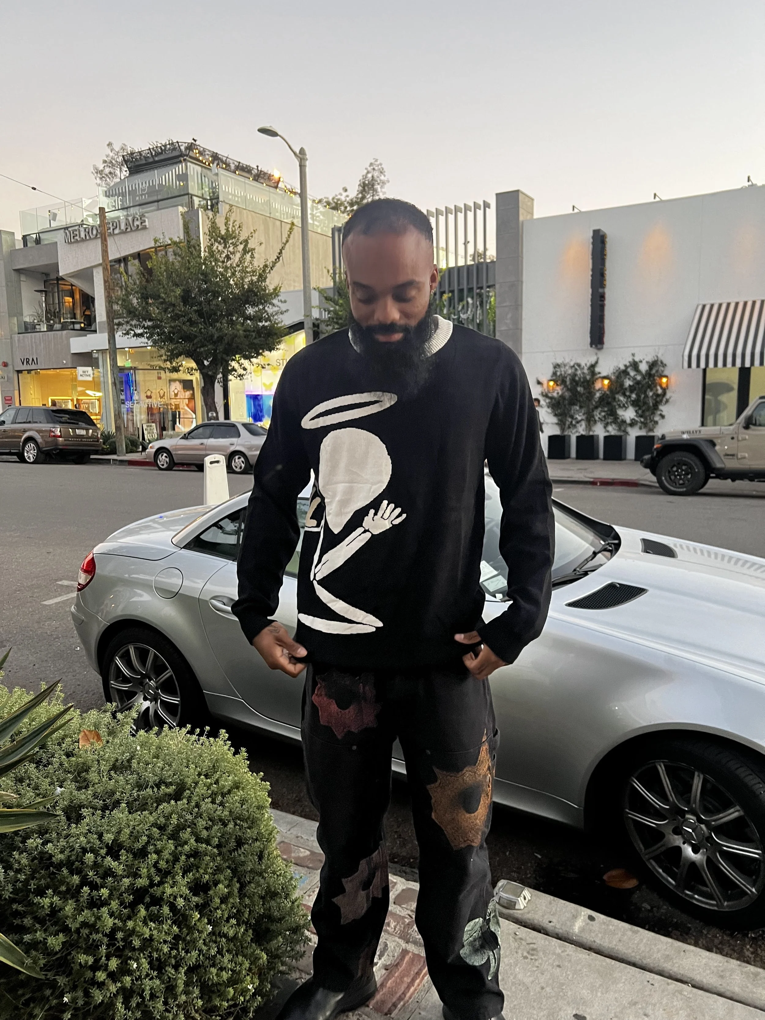 A man with a beard and short hair standing outdoors on a city street with parked cars behind him, wearing a black sweater with a large white skull design and multicolored patched pants, during dusk.