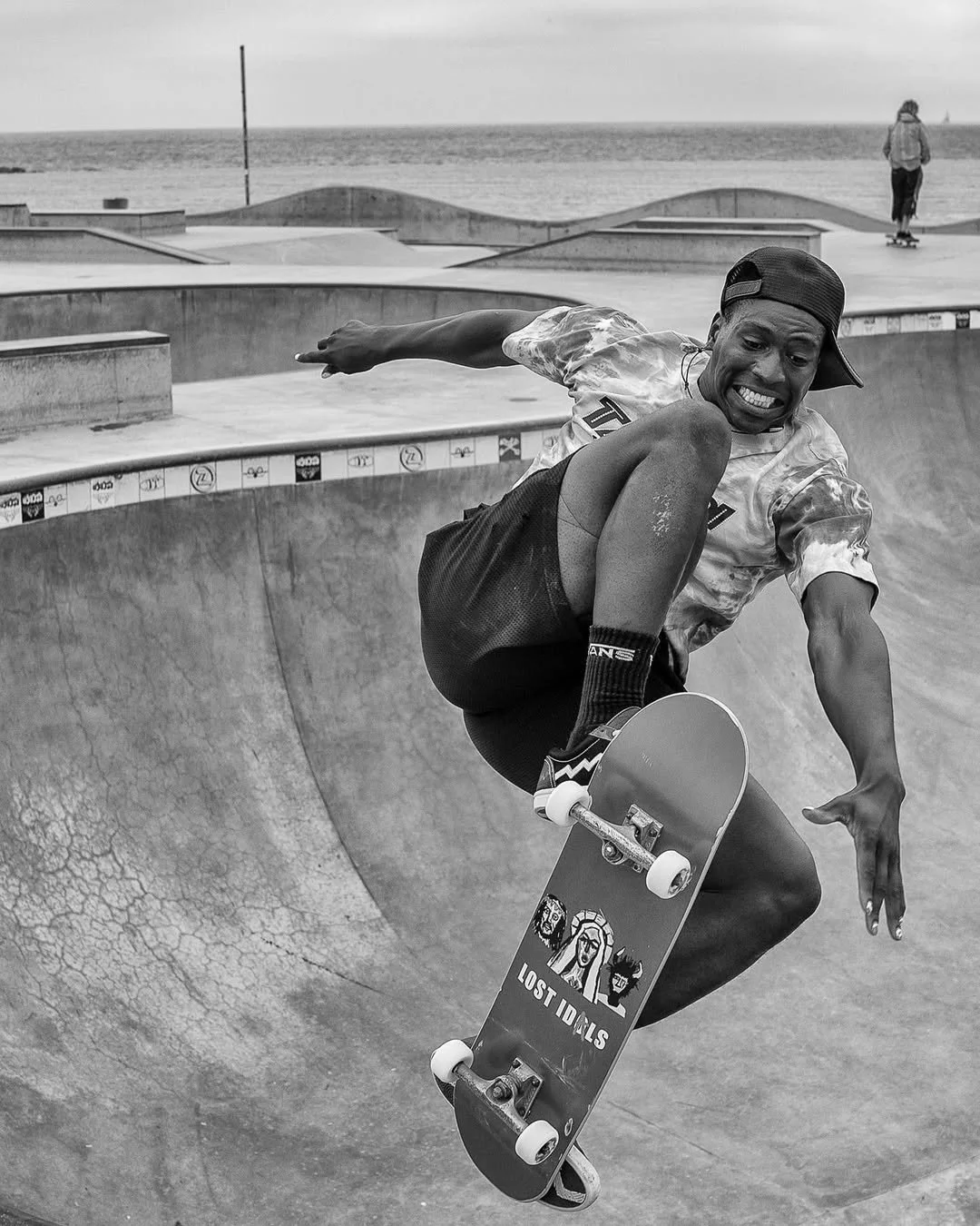 A person skateboarding inside a skatepark, mid-air with a beach and ocean in the background, black and white photo.