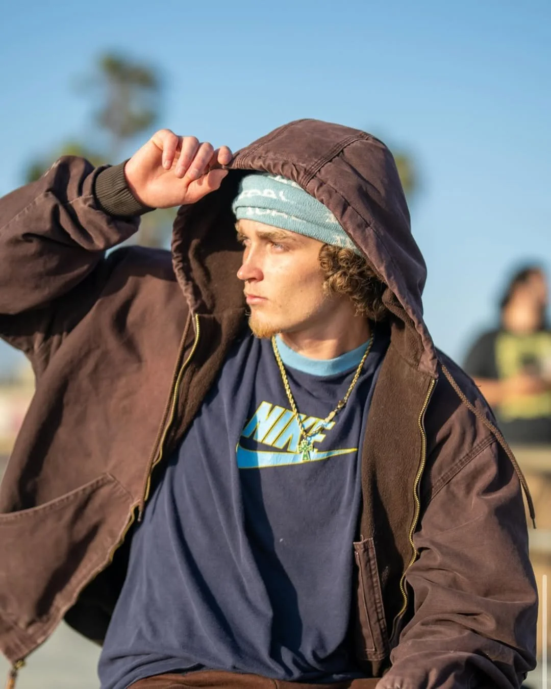 Young man wearing a brown hoodie, blue Nike t-shirt, and a light blue beanie, shielding his eyes from the sun with his hand, outdoors with a blurred background.