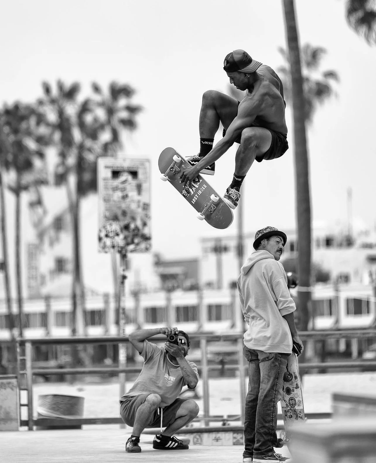 A shirtless skateboarder wearing a cap in mid-air performing a trick at a skatepark, witnessed by a young man with a skateboard and a photographer capturing the moment.