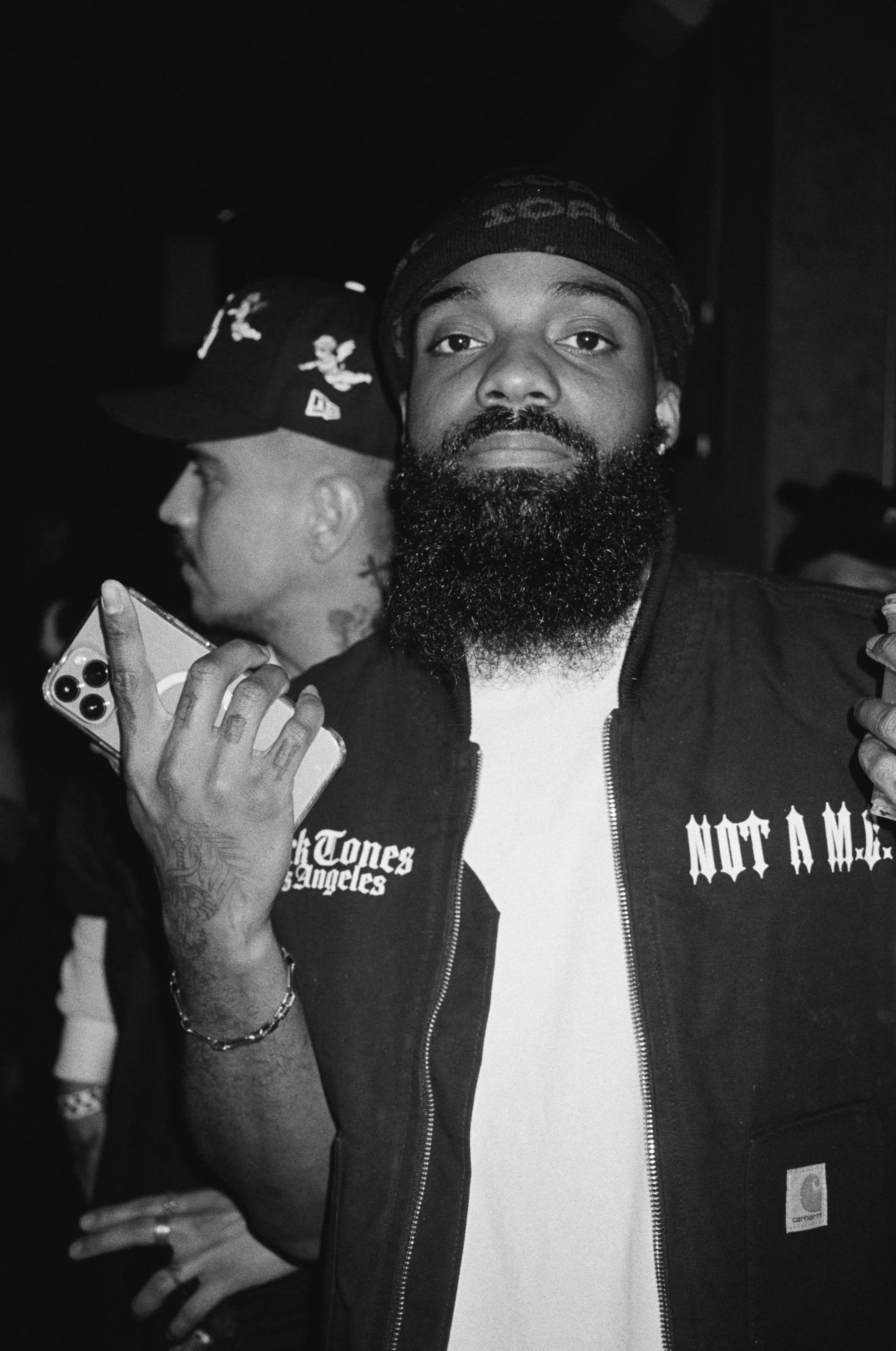 Black man with a full beard holding a smartphone, wearing a black baseball cap and a black jacket with patches, in a crowded setting.